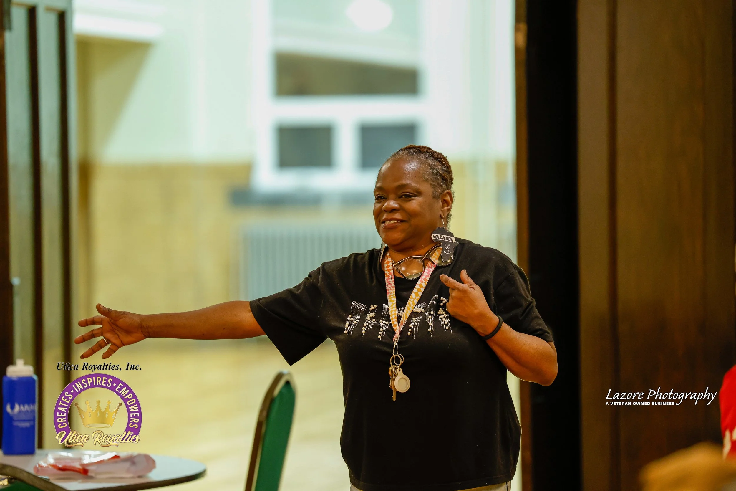 A woman with short hair, wearing a black T-shirt with white design, is smiling and gesturing with her right arm extended, in a room with yellow walls. She has a black wristband, a lanyard with keys, and large earrings shaped like the Wakanda flag.