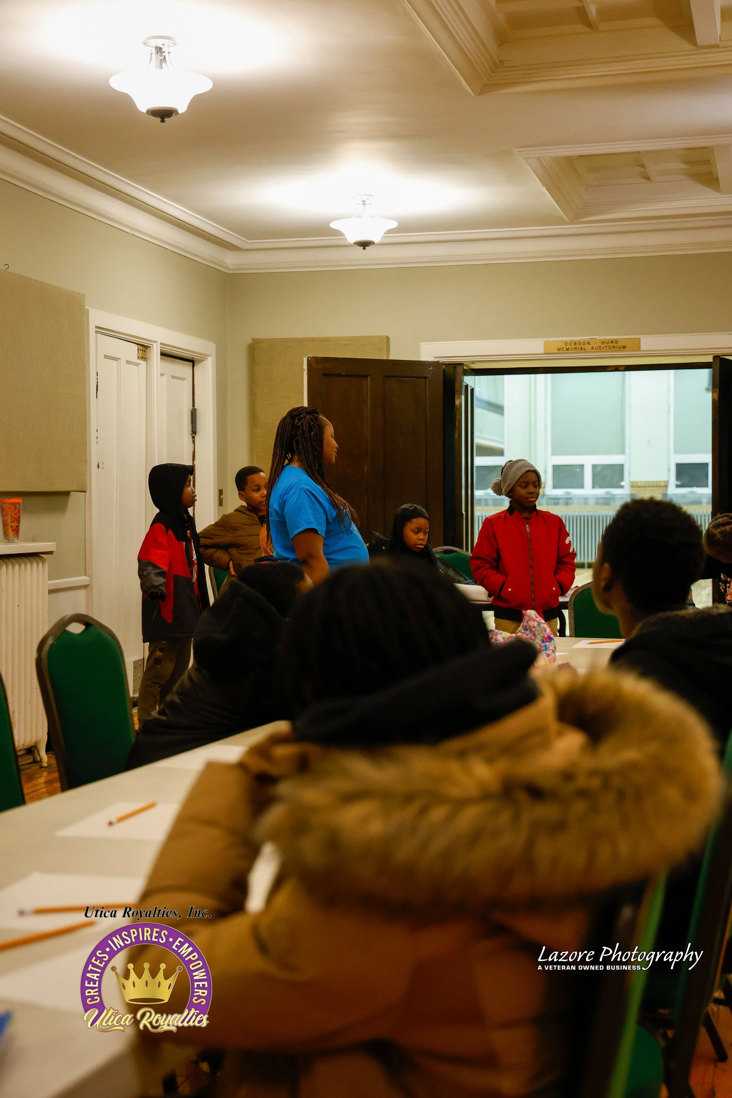 A group of children standing in front of a room, speaking or presenting while others sit at tables listening. The room has a high ceiling with lights, and an open door shows a building outside.