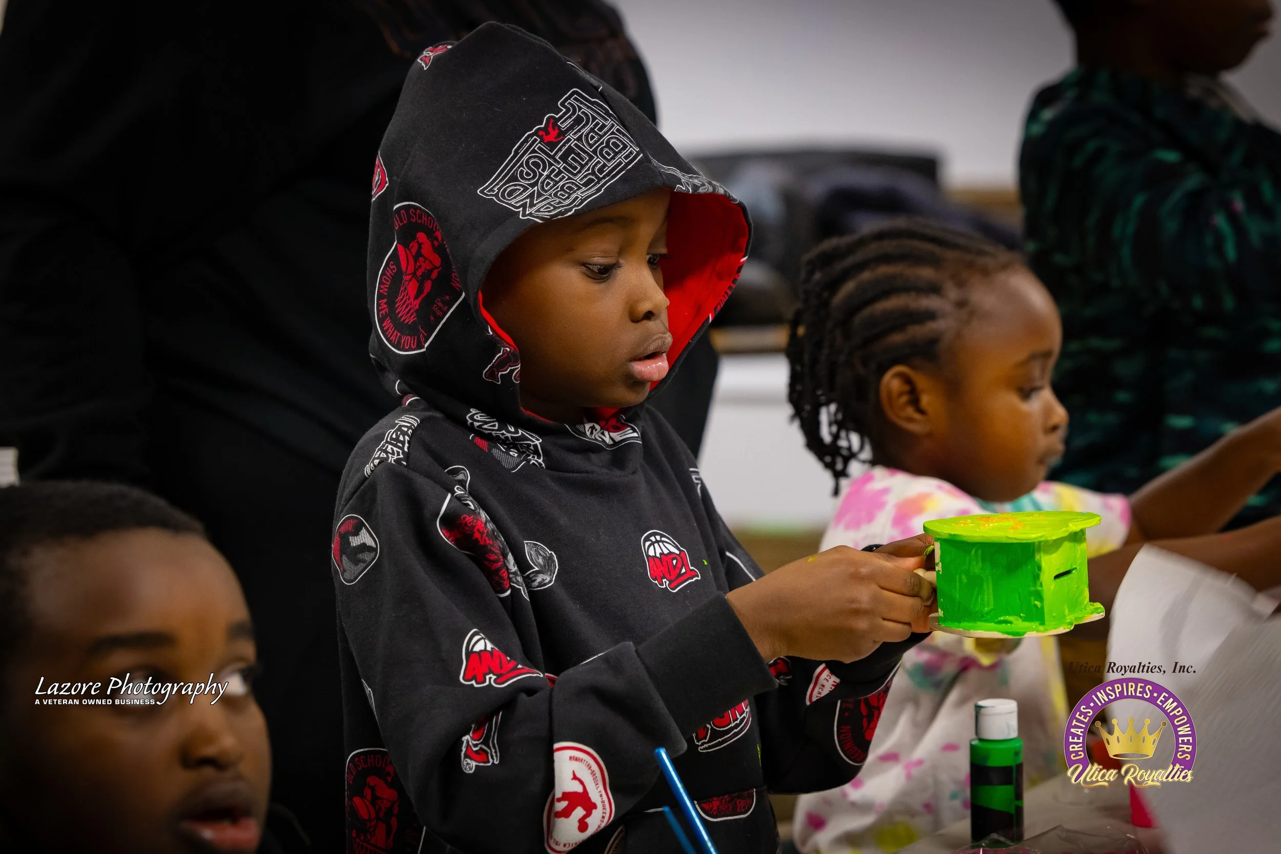 A young boy wearing a black hoodie with red and white graphics is holding a bright green paper plate or cup, looking at it with curiosity. Other children are around him, at a table, engaged in an activity.