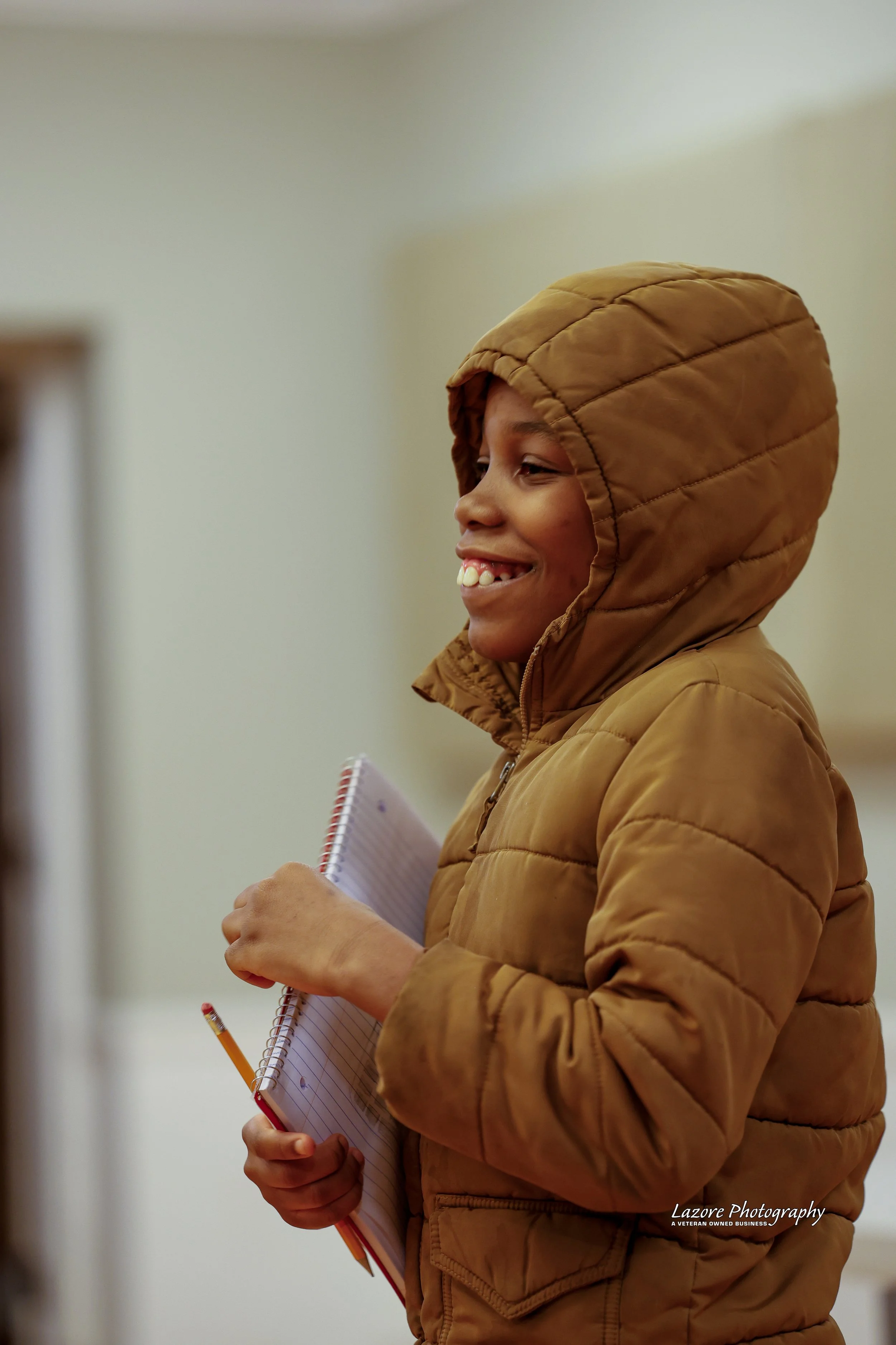 A young boy smiling while holding a notebook and pencil, wearing a brown hooded jacket.
