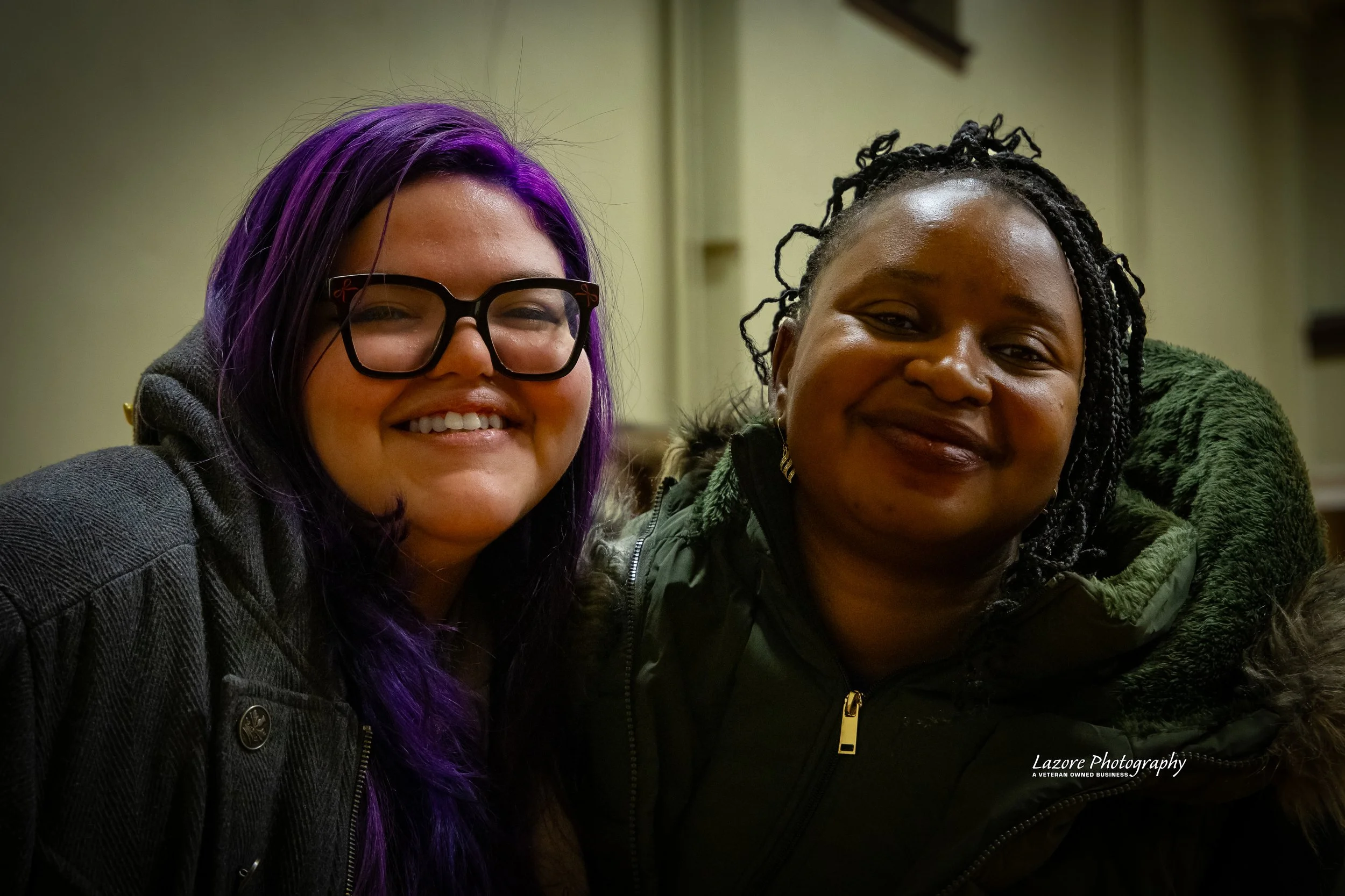 Two women smiling closely at the camera indoors, one with purple hair and glasses, the other with black braided hair, both wearing dark jackets.