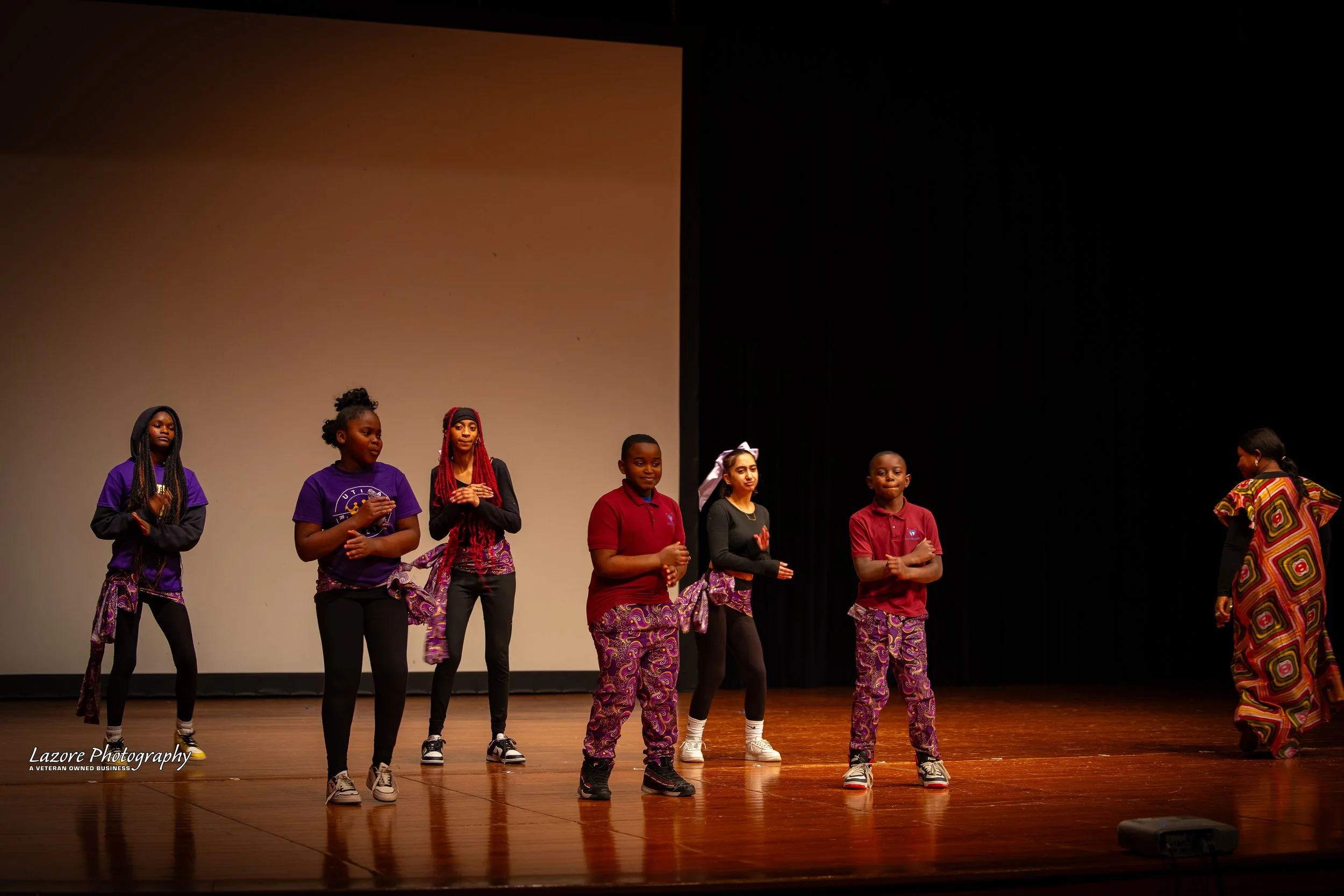 Children on stage performing in colorful outfits with one adult woman in a patterned dress.