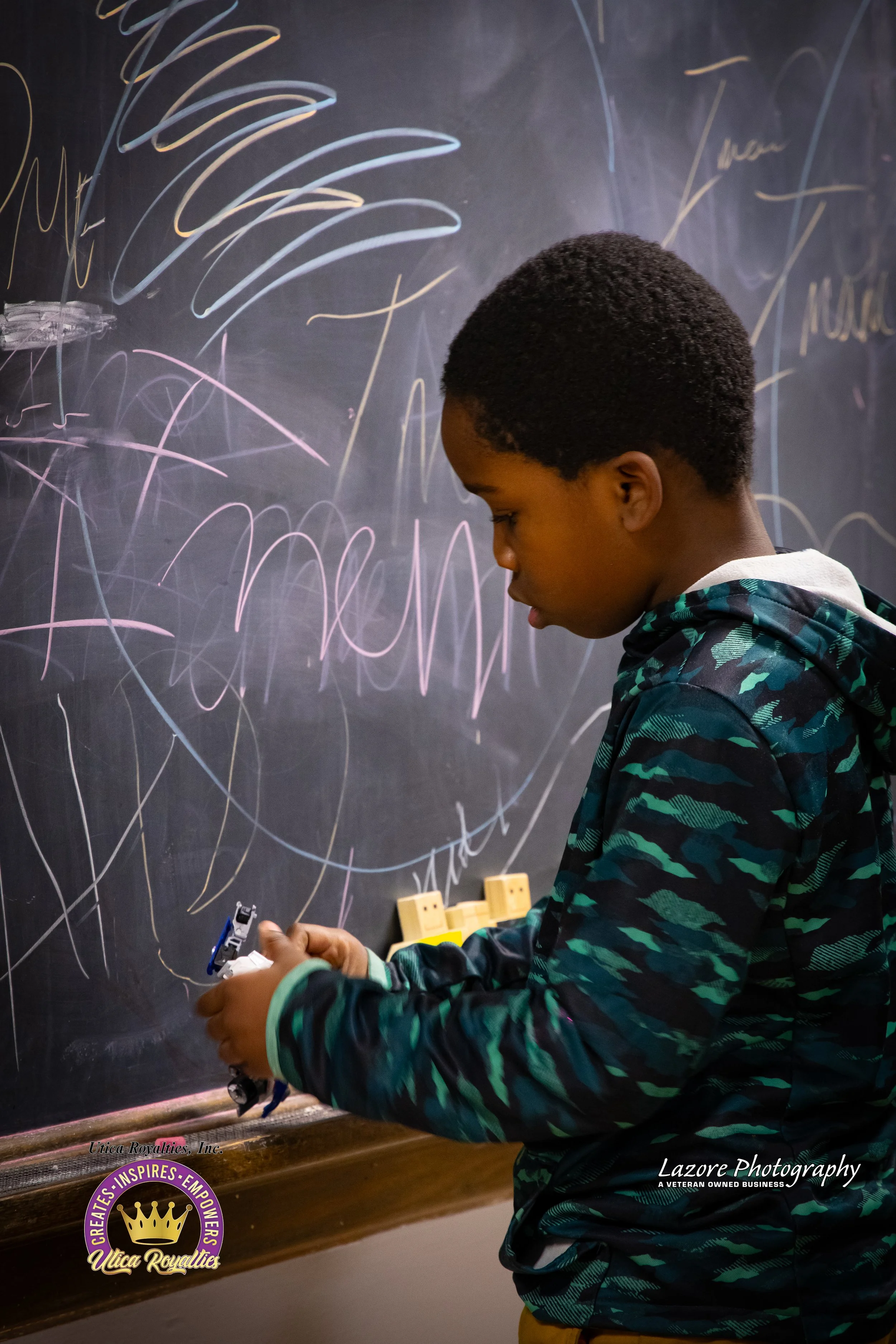 A young boy wearing a camouflage jacket writes on a black chalkboard with colorful chalk, and the words 'team' and 'mama' are visible among the scribbles.