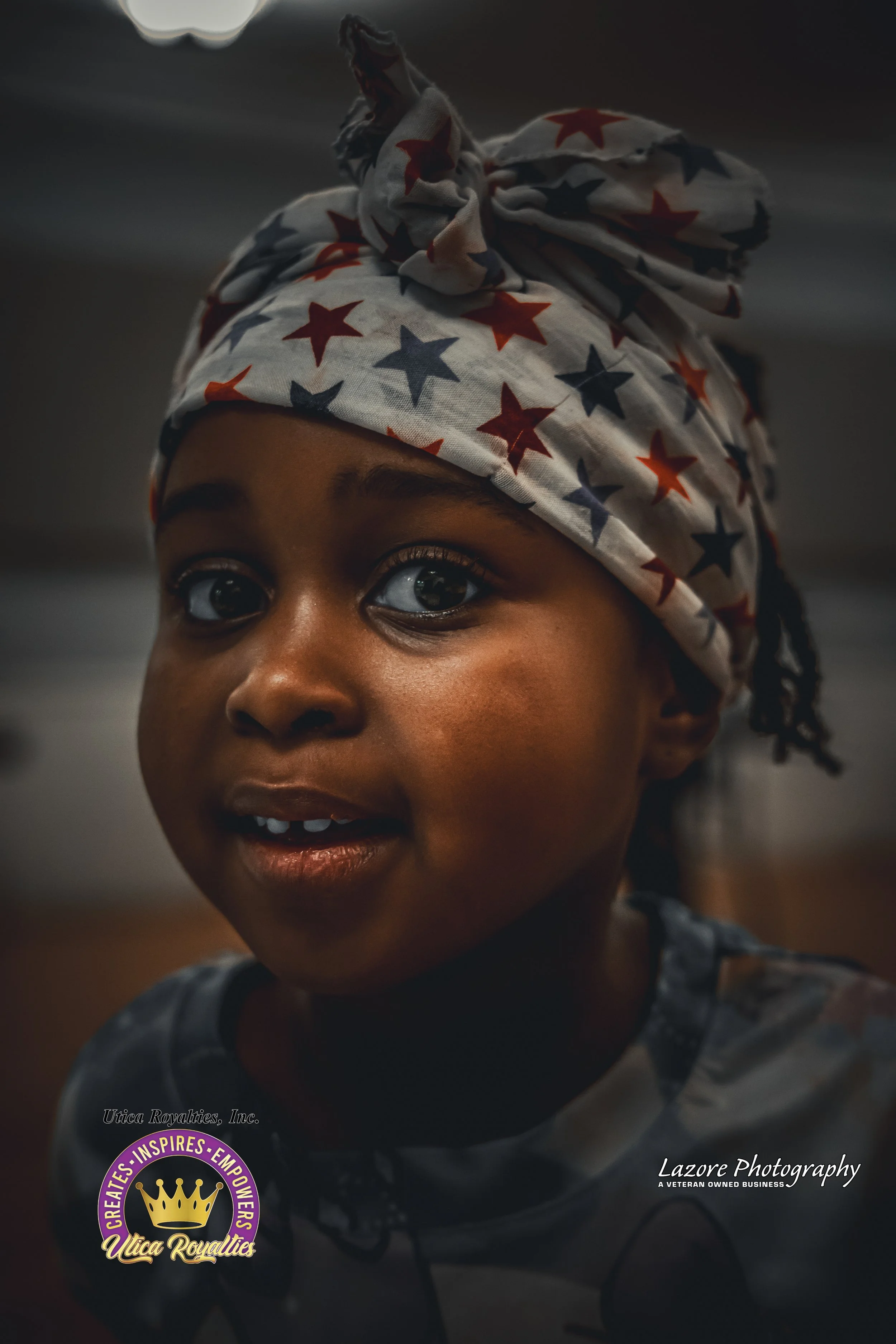 Close-up of a young girl with dark skin wearing a star-spangled headscarf and a dark patterned shirt, smiling indoors.