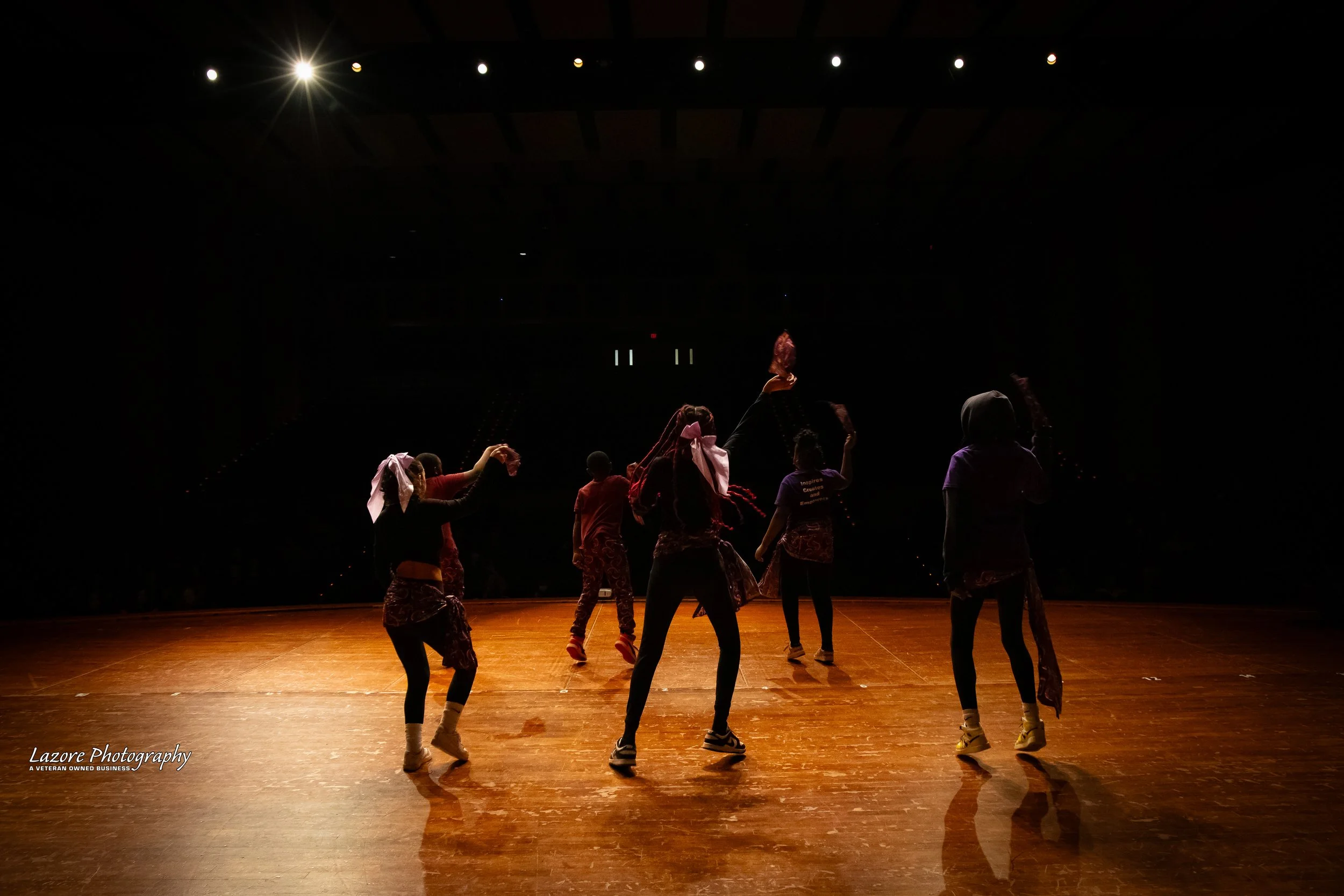 Group of five young dancers practicing on an empty stage with a wooden floor, under bright stage lights, with dark background.