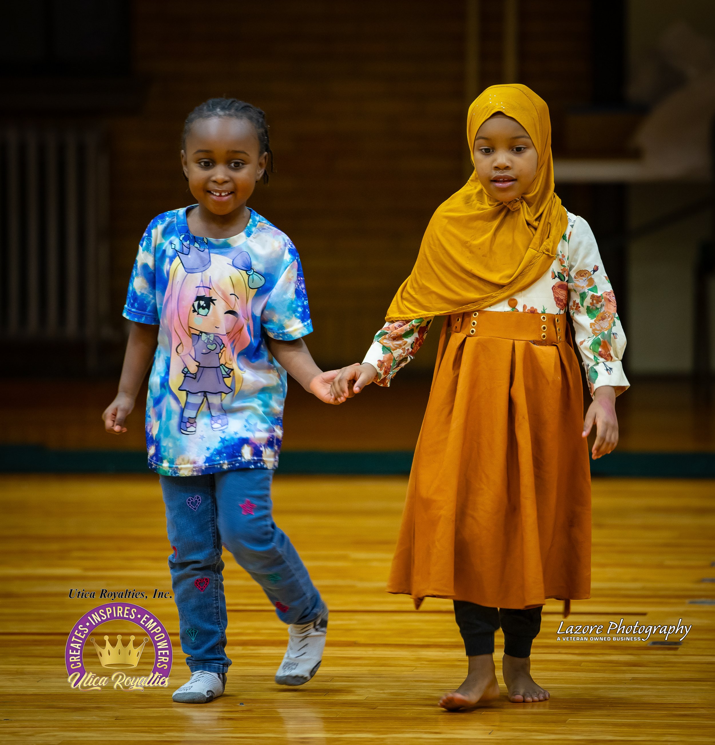 Two young girls holding hands and walking on a wooden floor, smiling and engaging in a dance or performance. One girl is dressed in a colorful t-shirt with a cartoon character, jeans, and sneakers. The other girl is wearing a mustard yellow hijab, a 