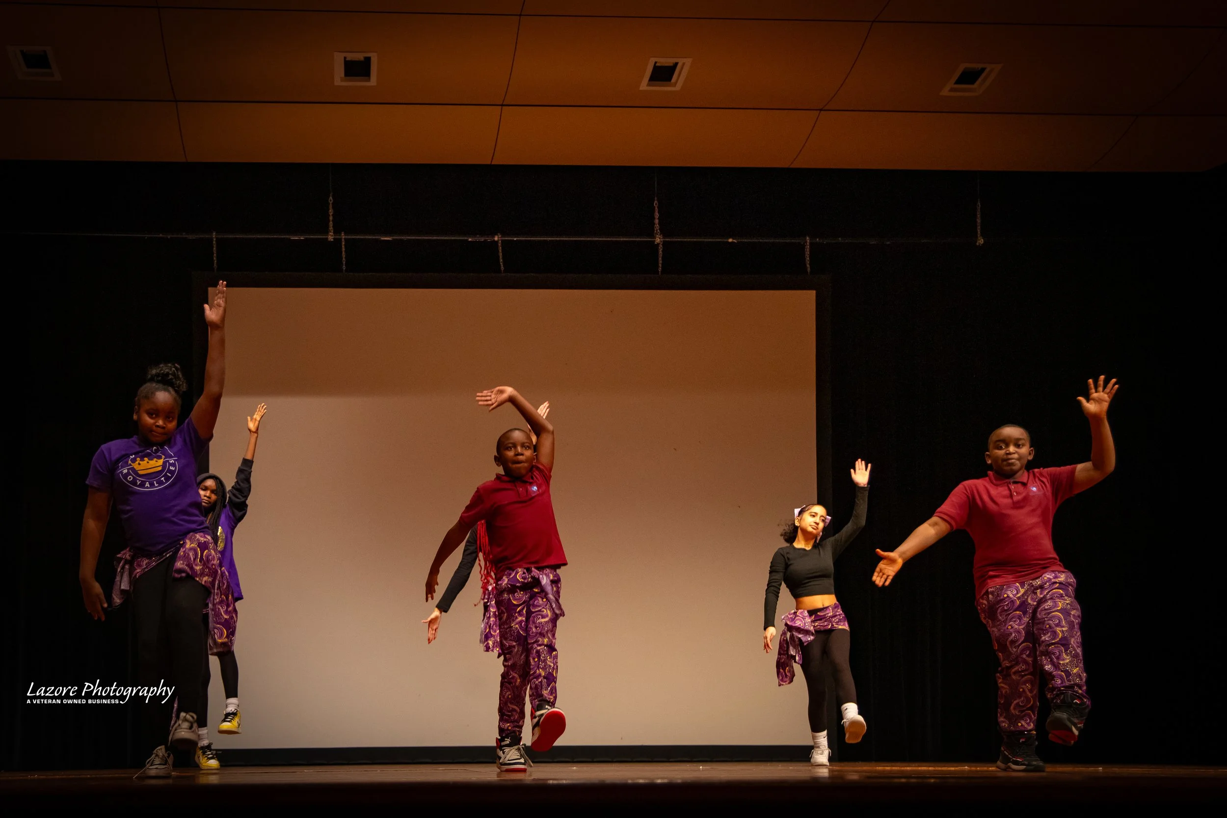Five children performing a dance routine on stage, wearing colorful outfits including purple, red, and black, with some raising their arms and moving in a coordinated manner.