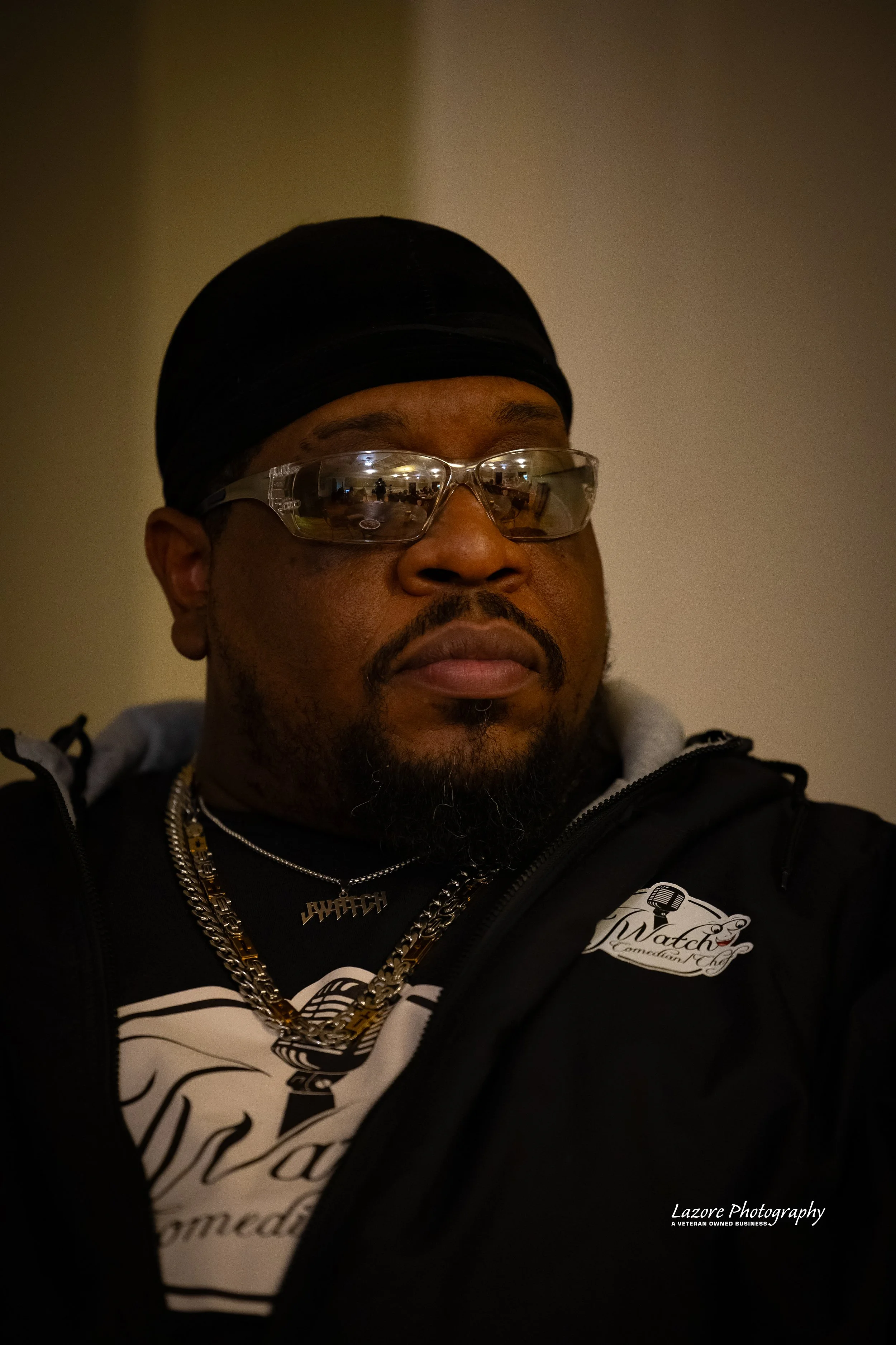 A man wearing a black durag, clear safety glasses, gold chains, a black jacket, and a T-shirt with the logo 'iWatch Commissary Chef'.