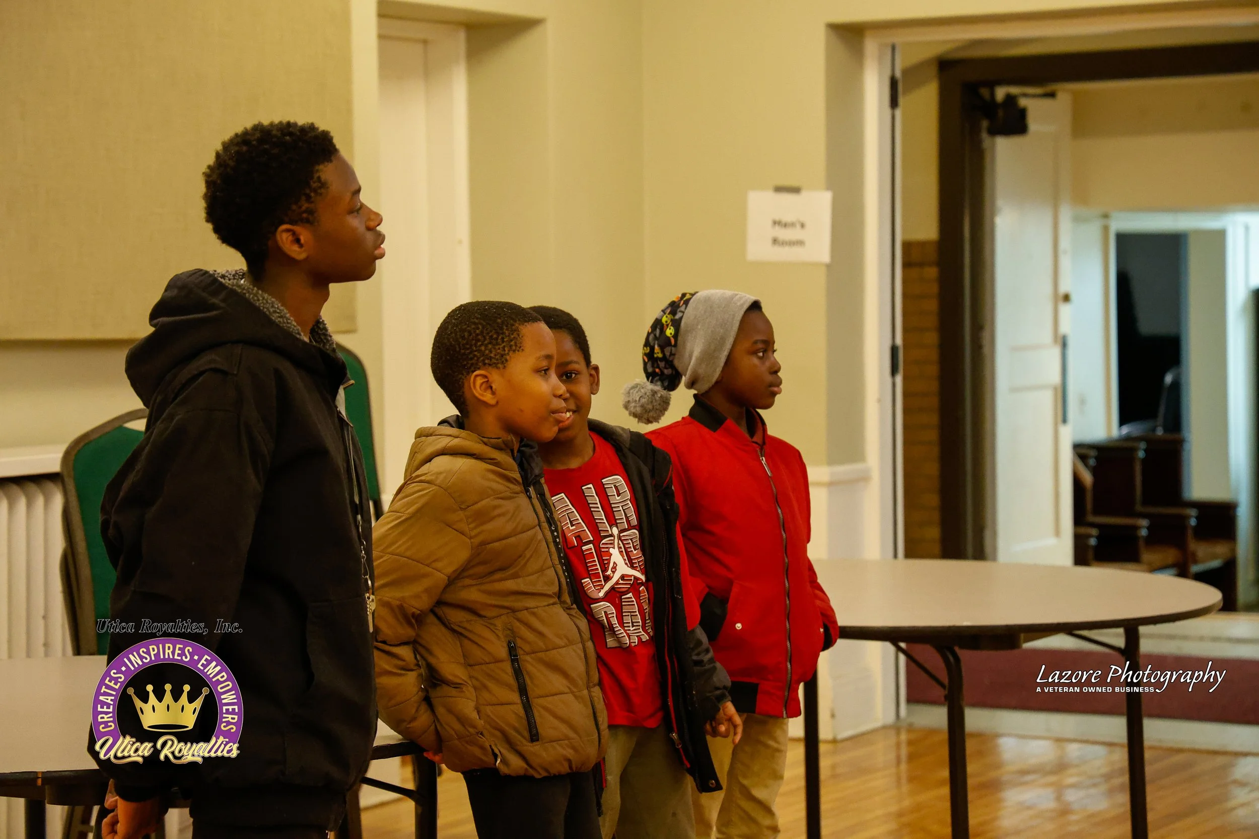 Four children standing in a row indoors, dressed warmly, with a light yellow wall and an open door in the background.