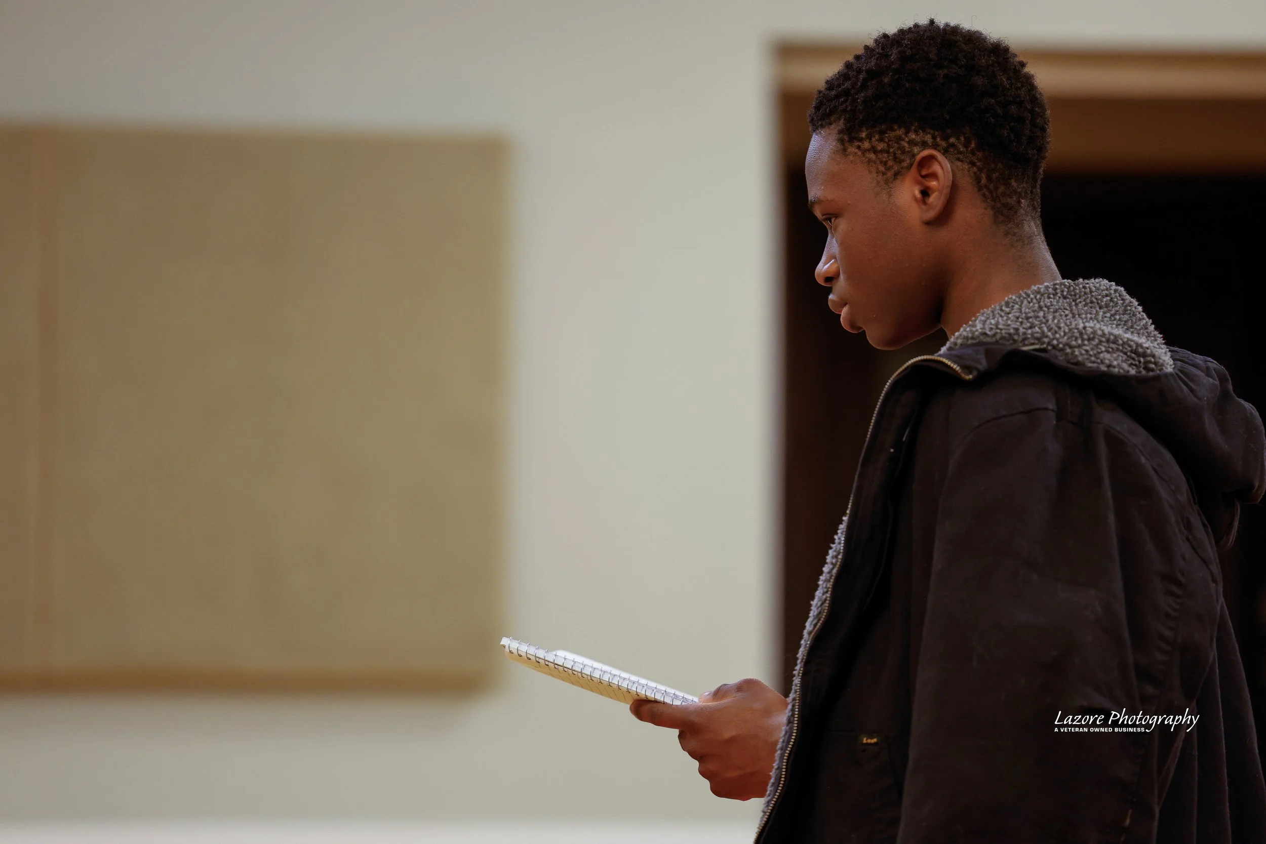 A young man standing in profile looking at a notepad in his hand, indoors with a blank wall and doorway in the background.