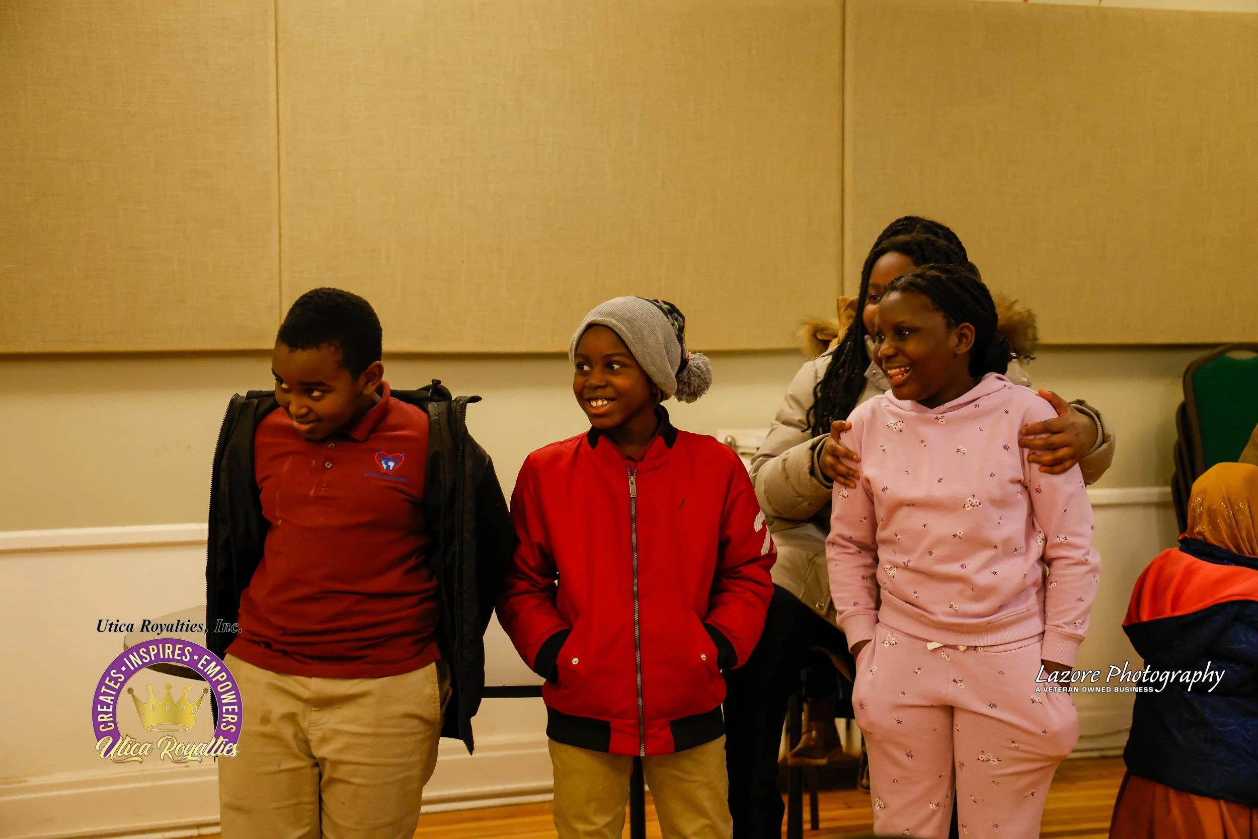 Three children standing indoors, smiling, with a woman embracing one of them in the background. The children are dressed in casual clothing, and the setting appears to be a community or event space.