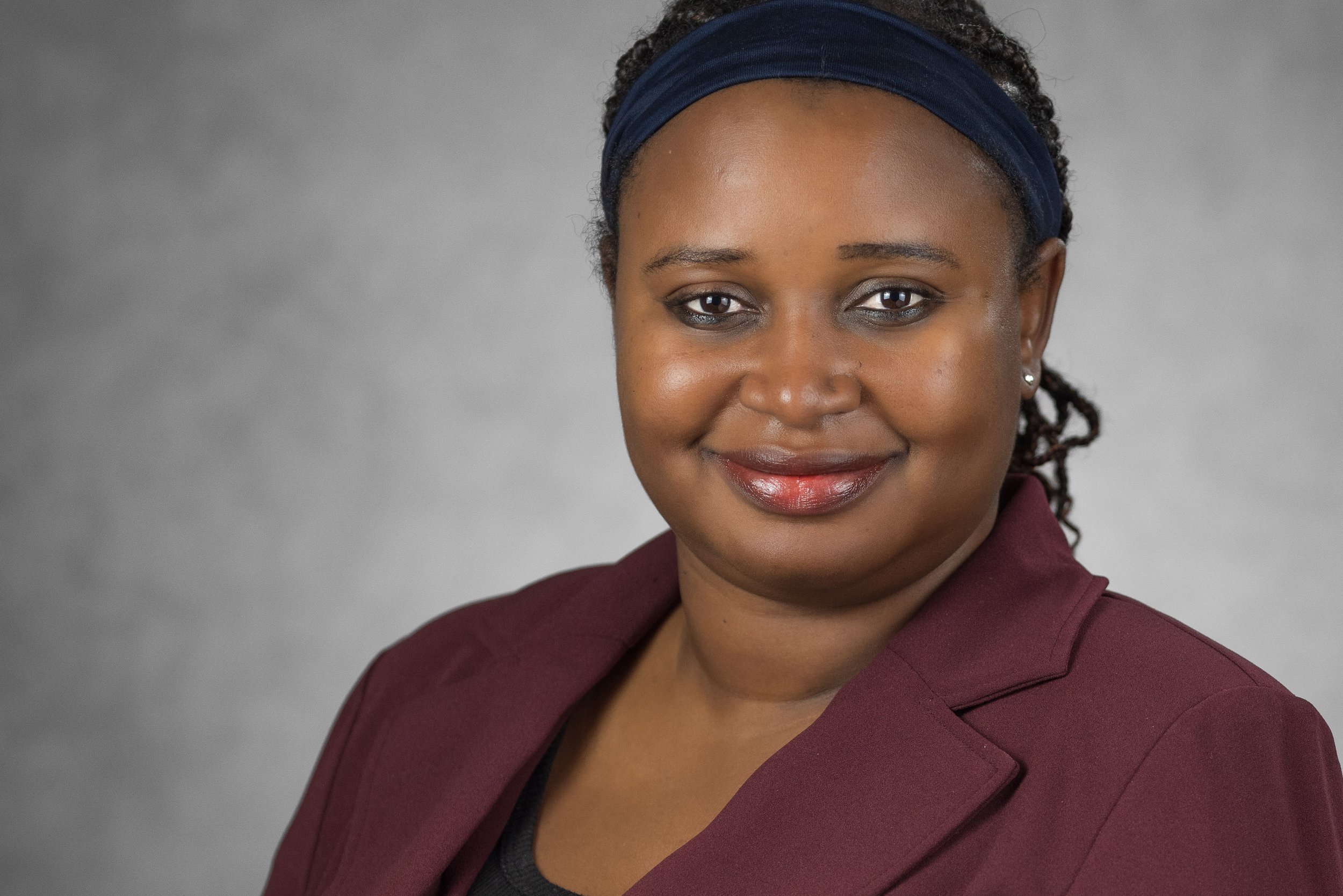 A woman wearing a bright blue headwrap decorated with flowers and gemstones, with bright red lipstick, silver earrings, and a black and white patterned blazer.