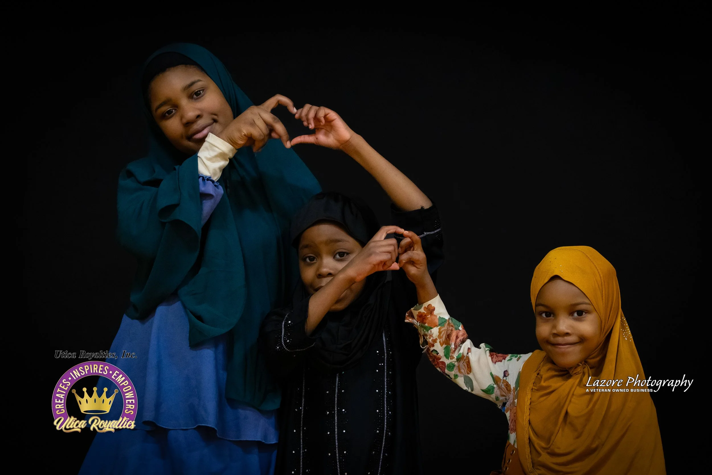 Three young girls wearing colorful headscarves and dresses, standing against a black background, making heart shapes with their hands.