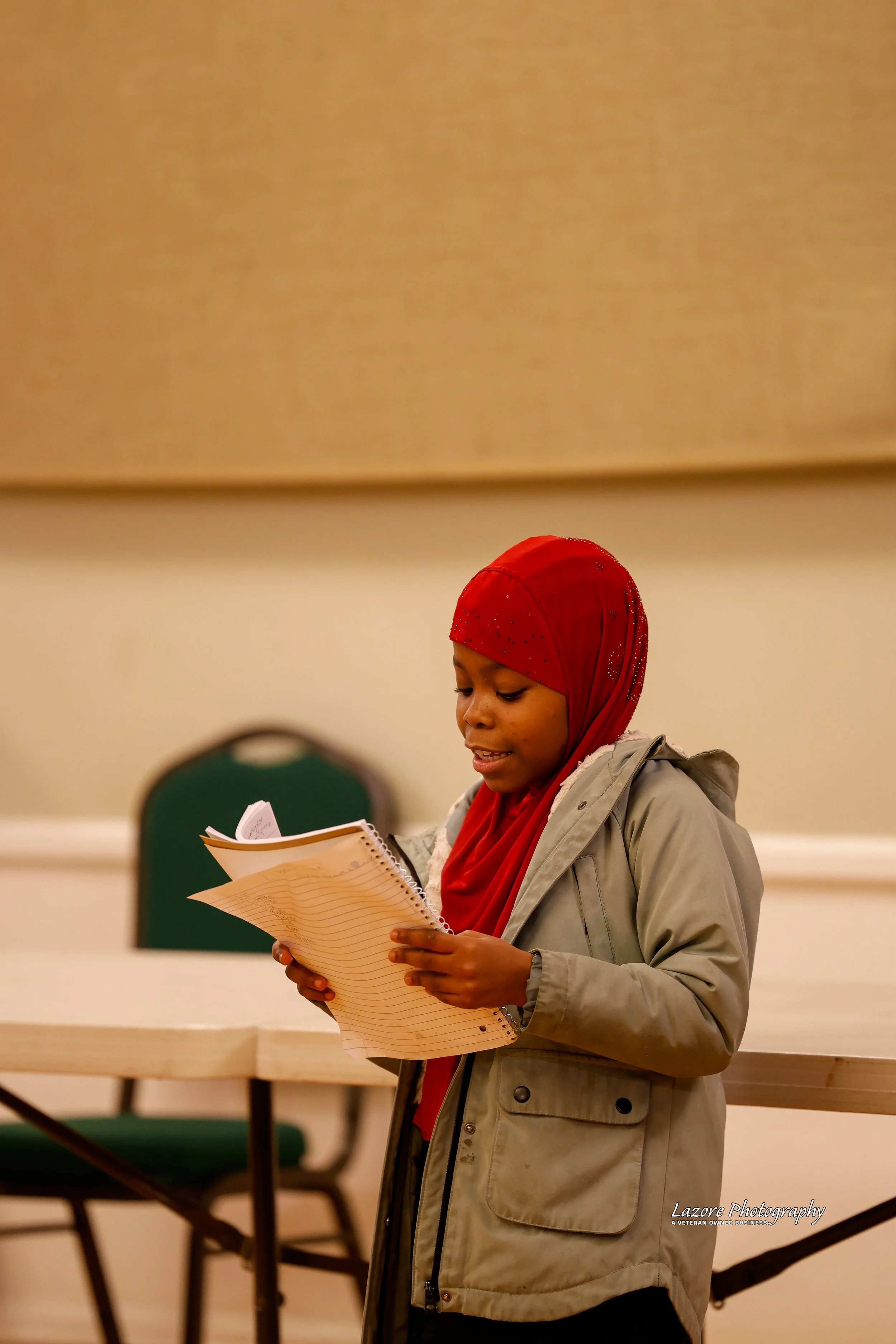 A young girl with a red headscarf and gray jacket reading from a spiral notebook in a room with beige walls and green chairs.