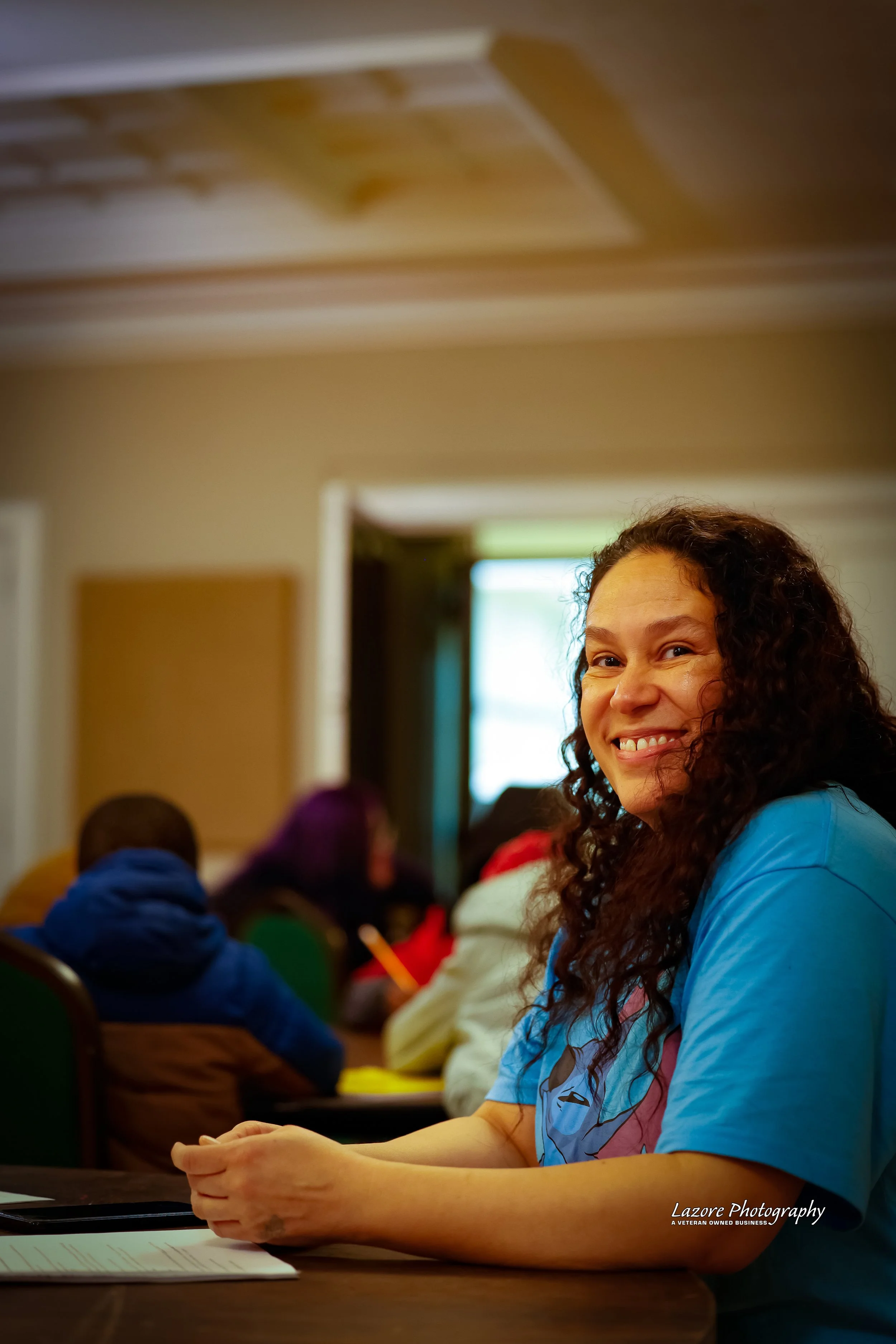 A smiling woman with curly hair wearing a blue shirt sitting at a table in a room with other people. The room has a ceiling with wooden paneling and a window in the background.