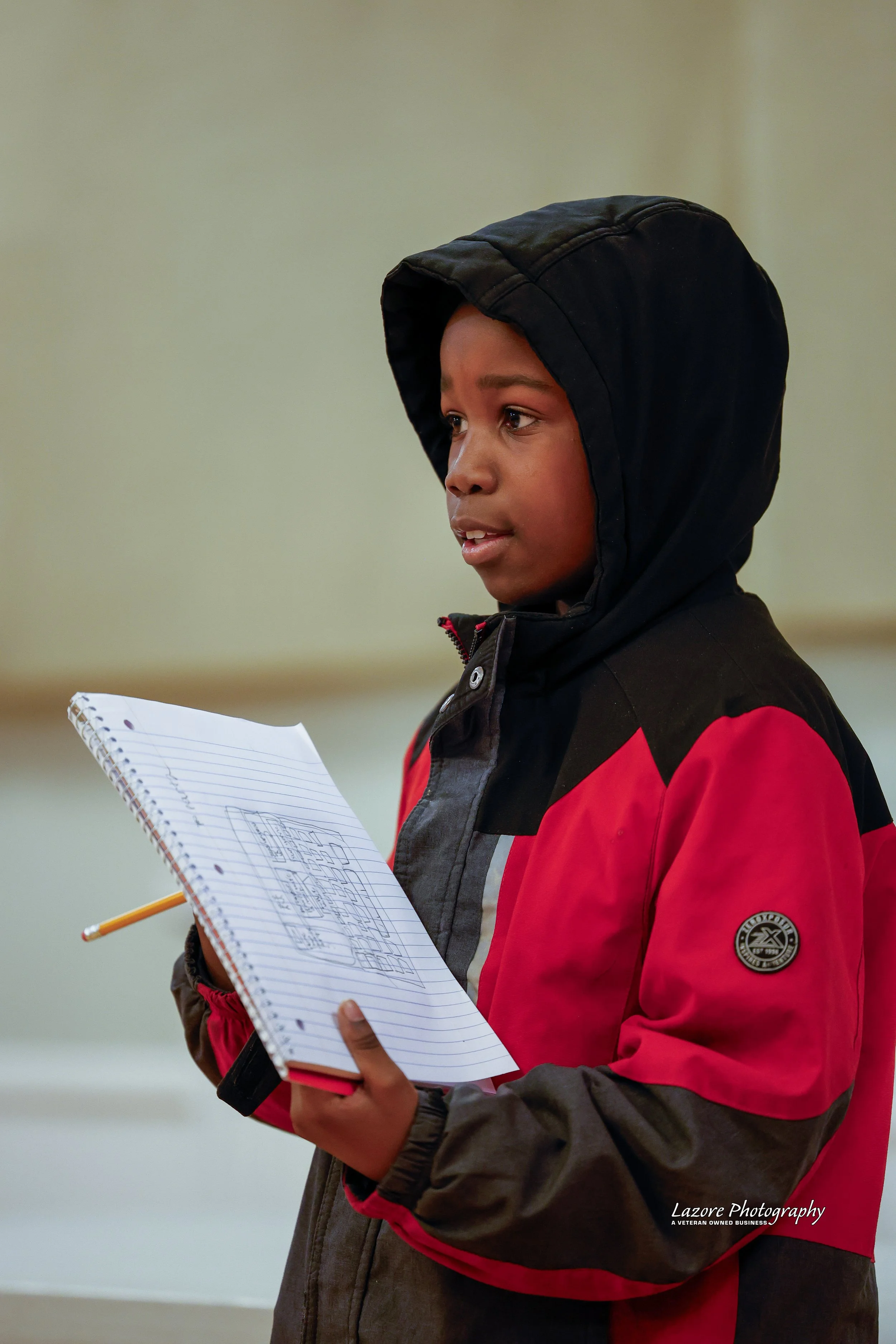 A young boy wearing a black and red jacket with a hood, holding a notebook and pencil, appears to be engaged in a discussion or presentation indoors.