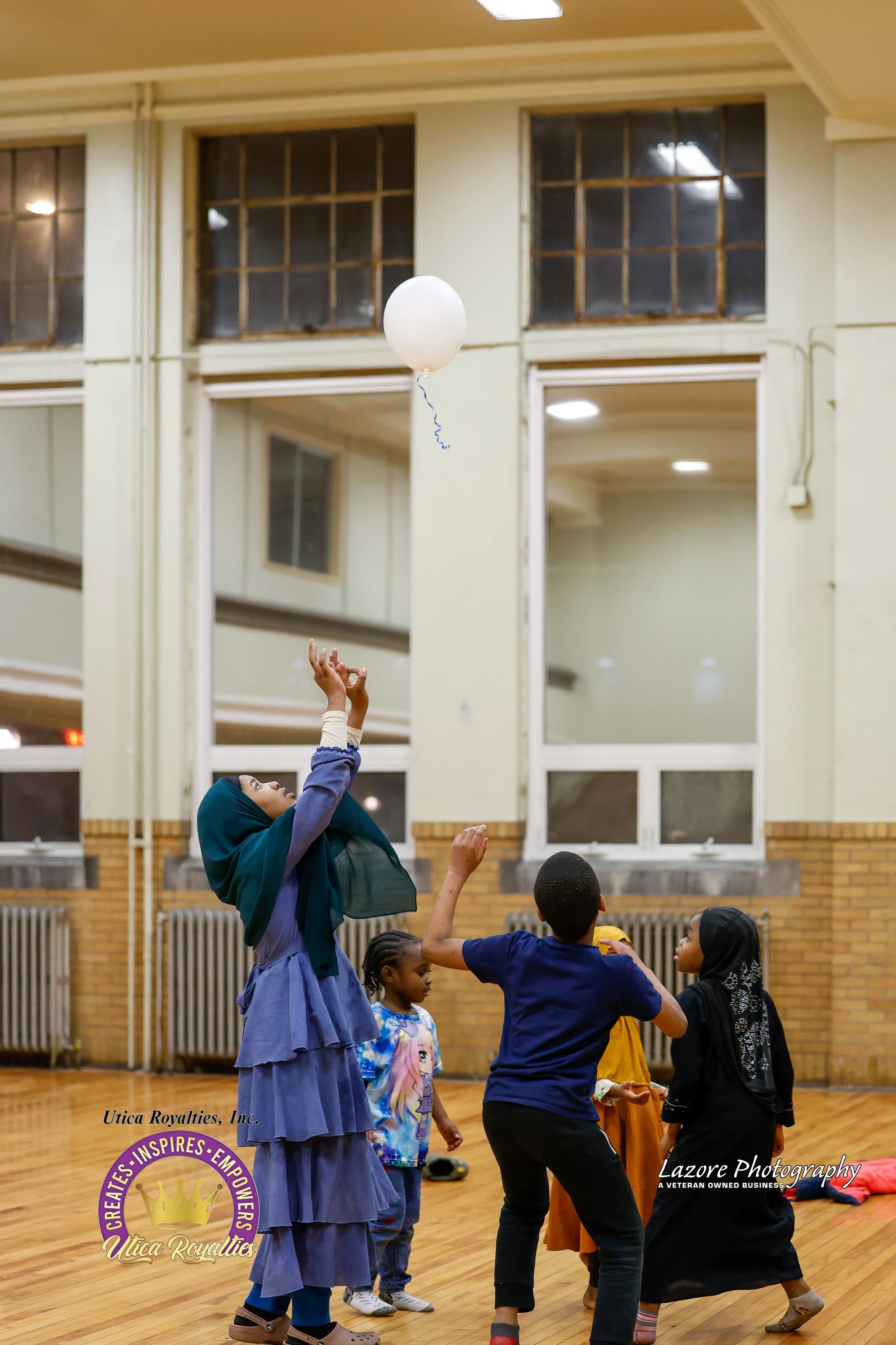 A group of children playing with a white balloon in a large indoor room with wooden floors and large windows.