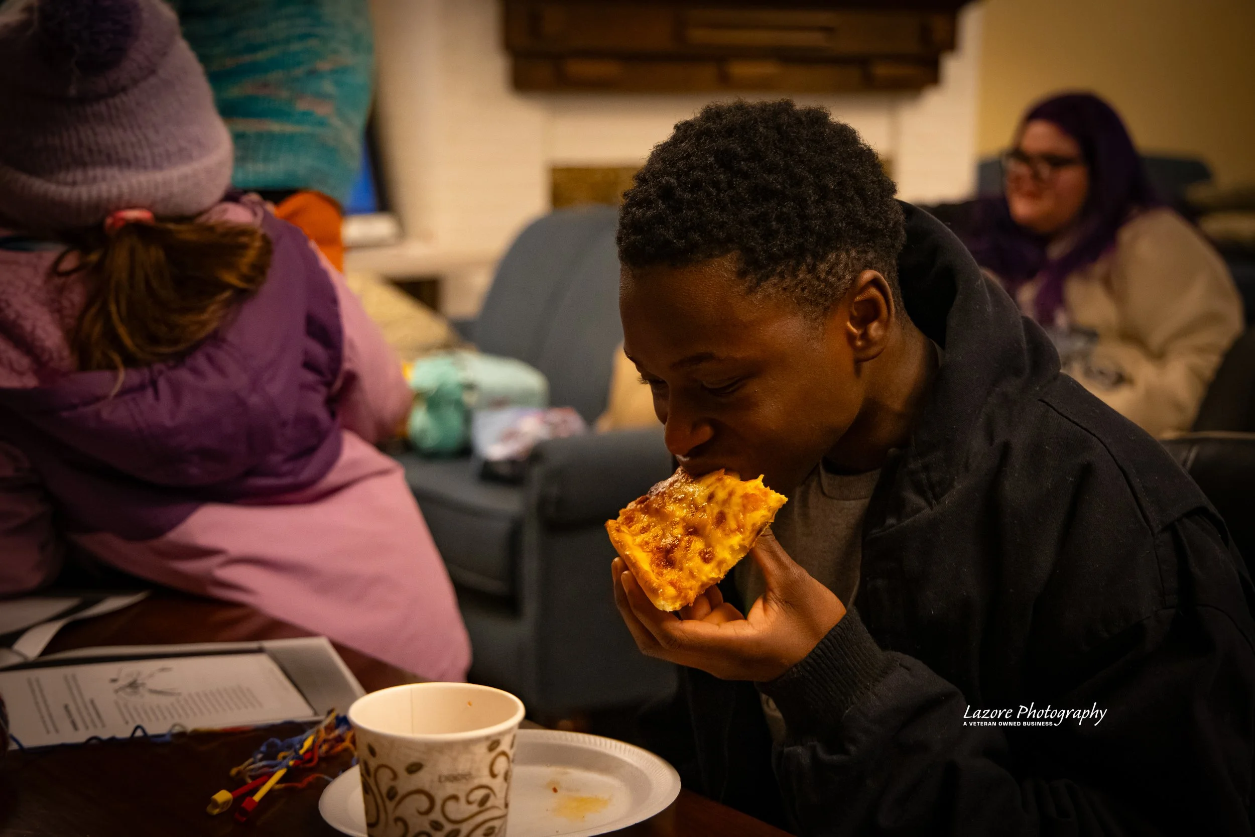 A young man is sitting at a table, biting into a slice of pizza, with a paper cup and some utensils on the table. In the background, there is a woman with purple hair sitting on a sofa and another person in a purple jacket, but their faces are not vi