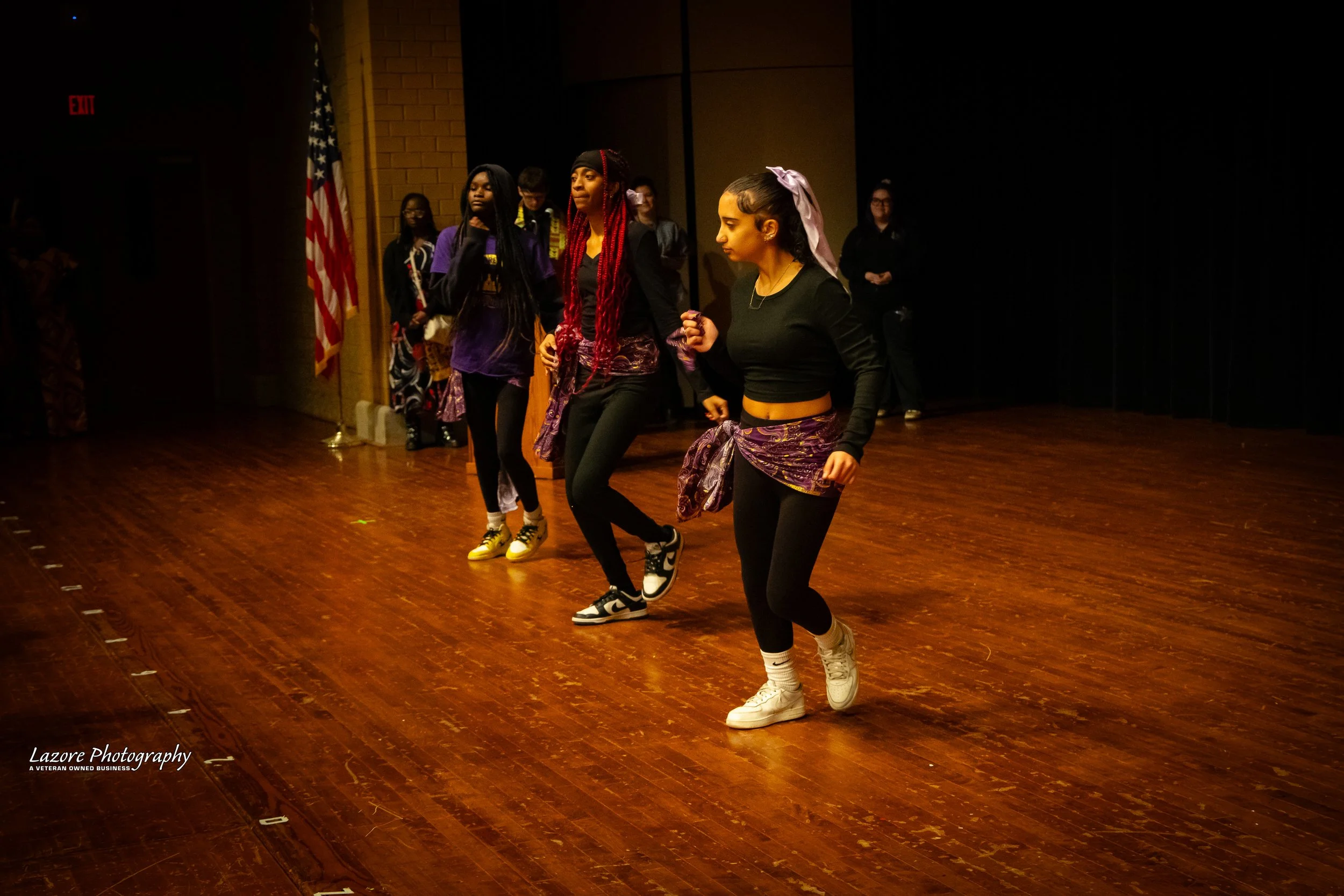 Young women dancing on stage with a wooden floor and a black curtain in the background, American flag visible on the left side.