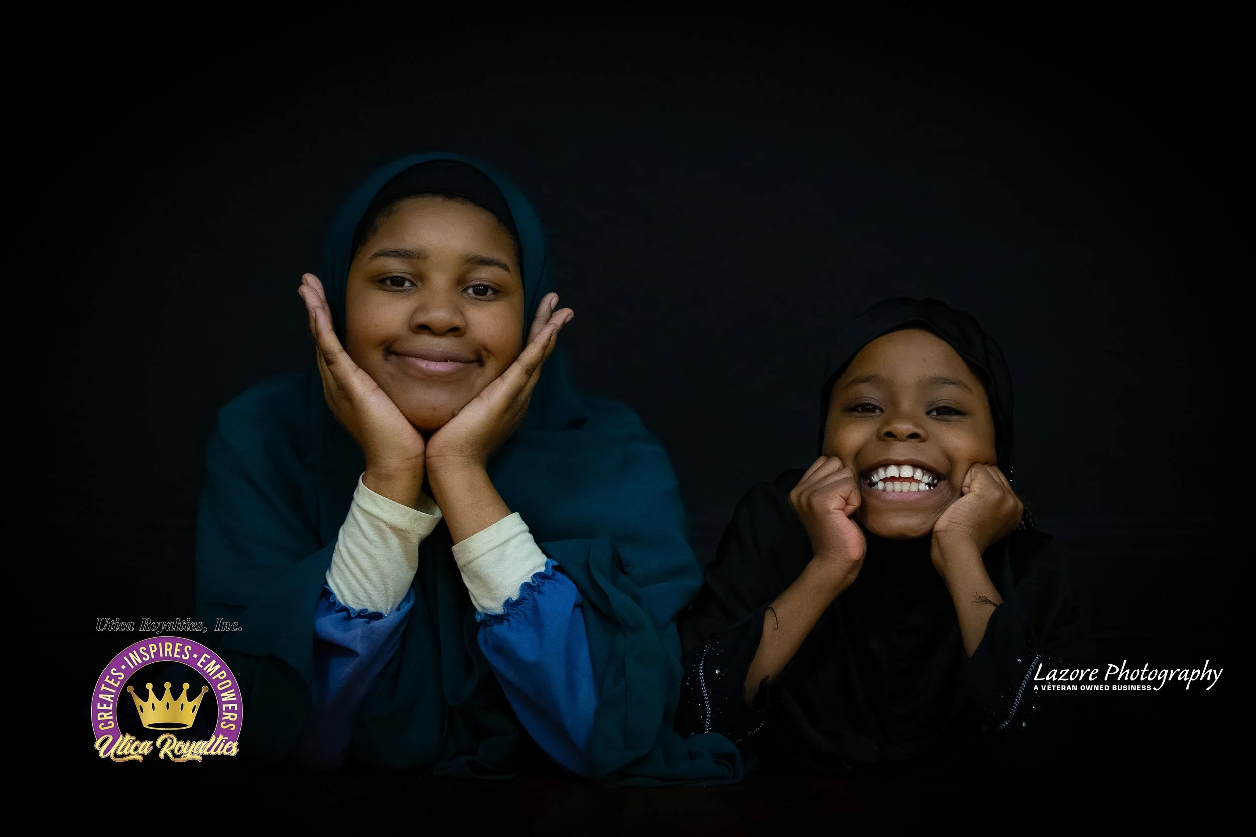 Two smiling young girls in hijabs, resting their chins on their hands against a black background.