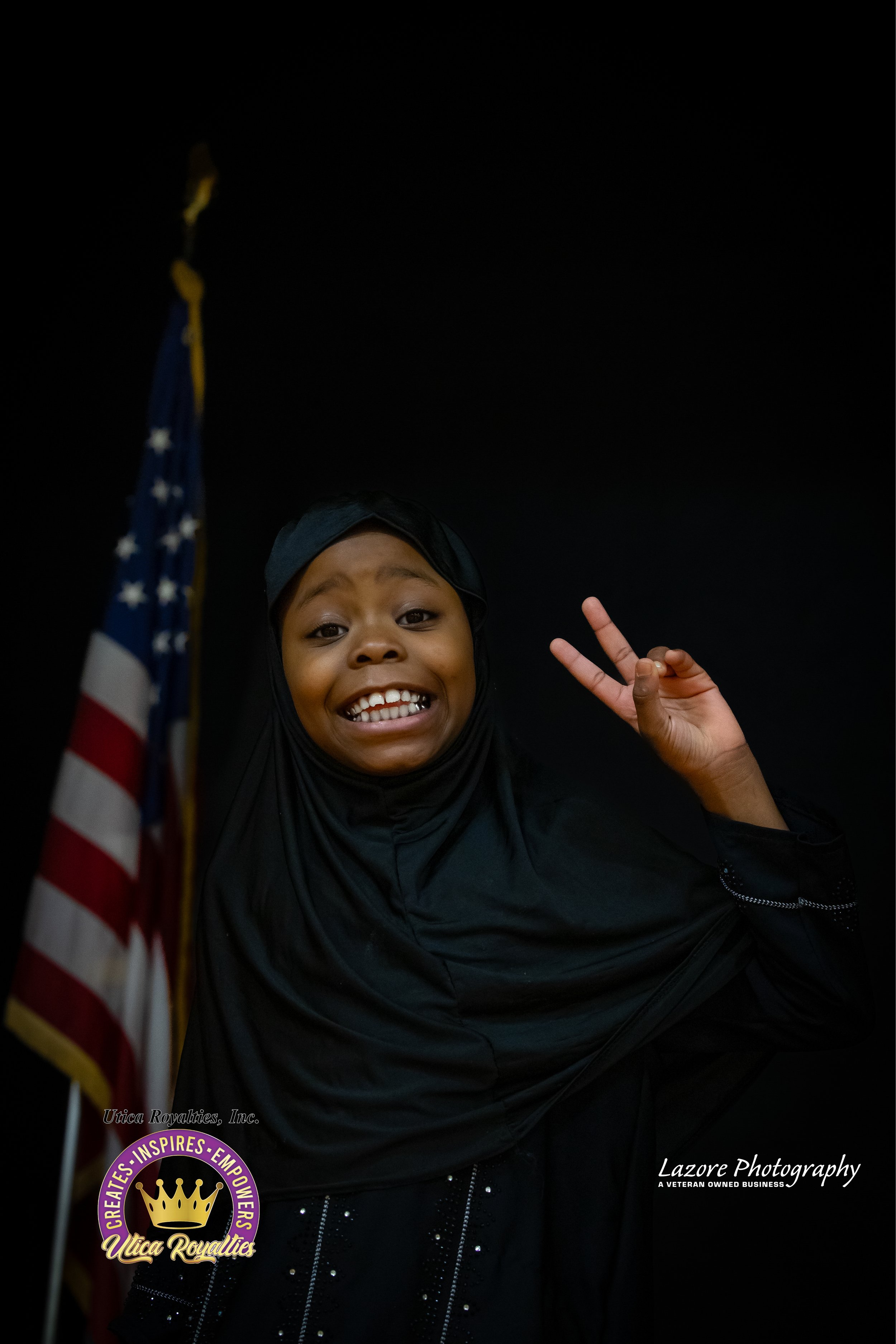 Young girl wearing a black hijab making a peace sign and smiling, standing beside an American flag, against a plain dark background.