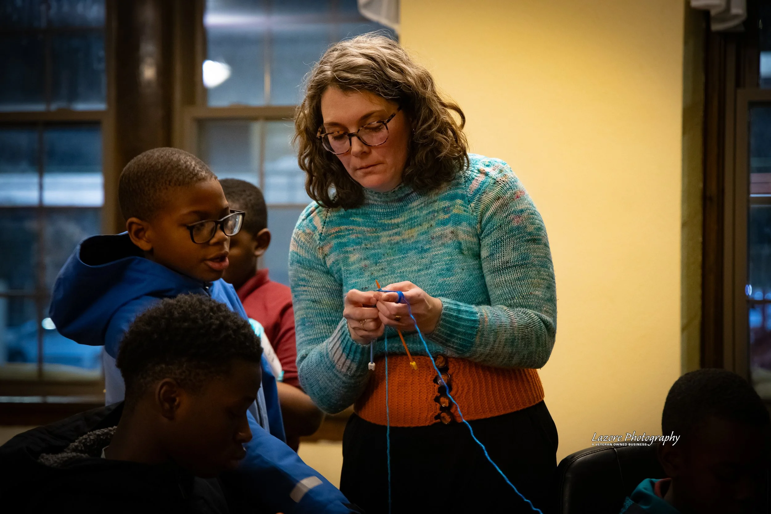 A woman teaching knitting to a group of children in a room with large windows and wooden frames.