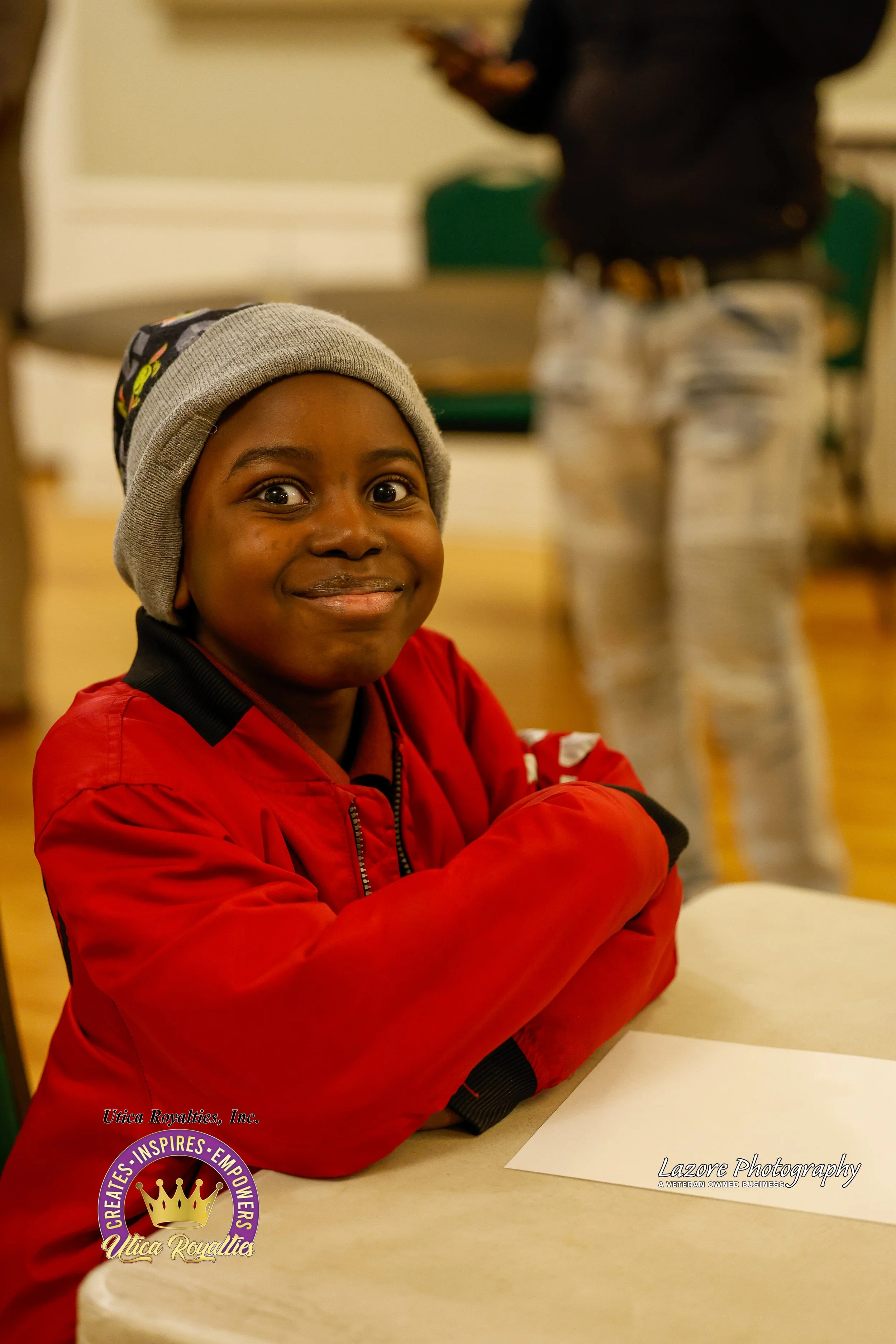 A young boy wearing a gray beanie and red jacket sitting at a table with a big smile, with other people in the background.