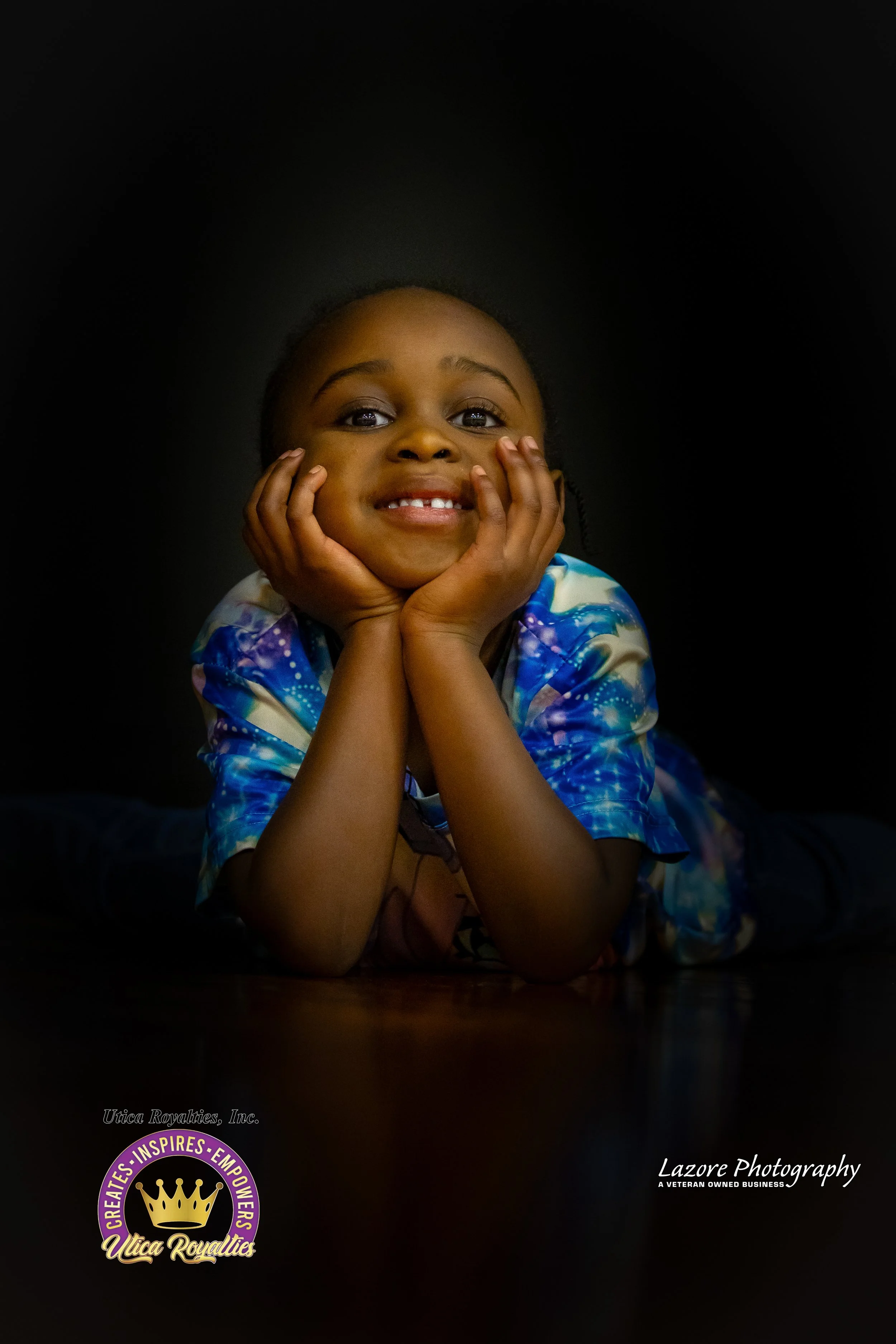 Young girl lying on the floor, resting her face in her hands, smiling at the camera, wearing a colorful tie-dye shirt, with a dark background.