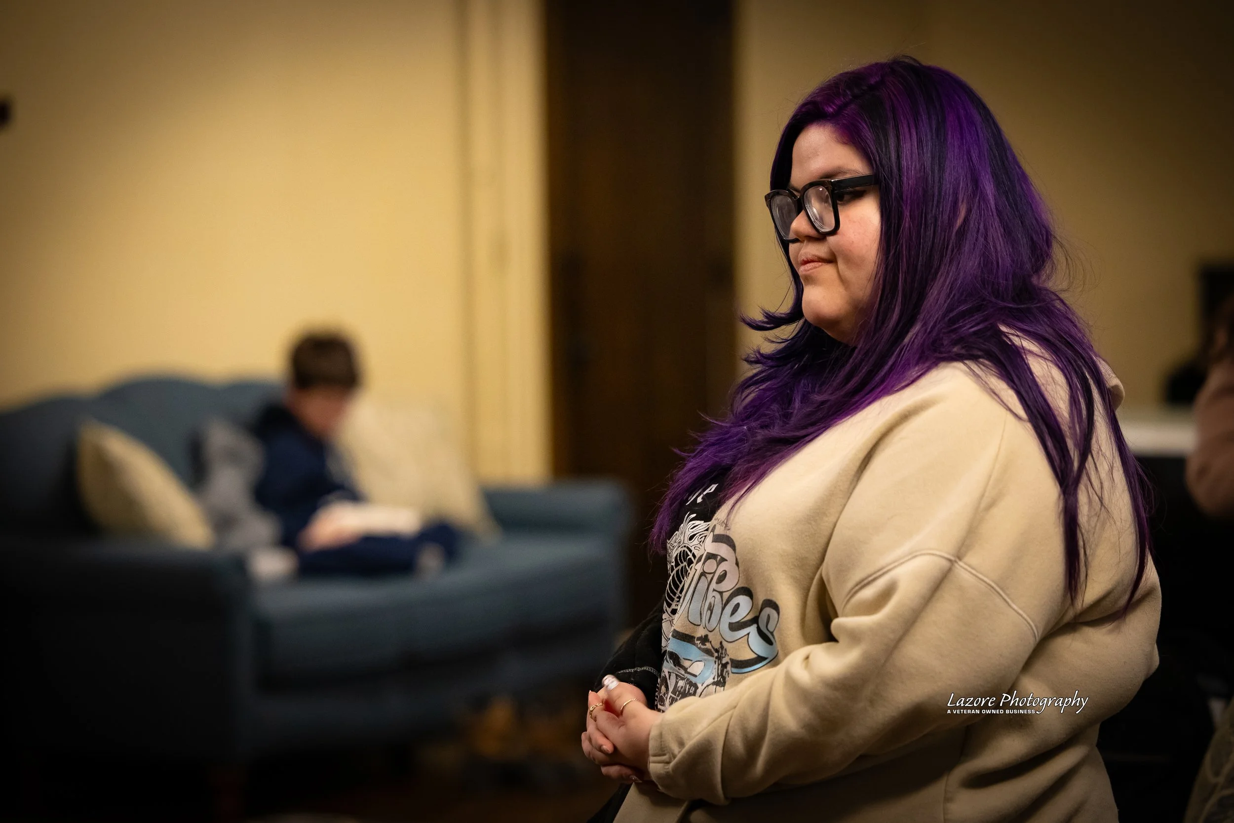 A woman with purple hair and glasses stands in profile, eyes closed, in a warmly lit room. A boy reading a book is blurred in the background on a dark teal couch.