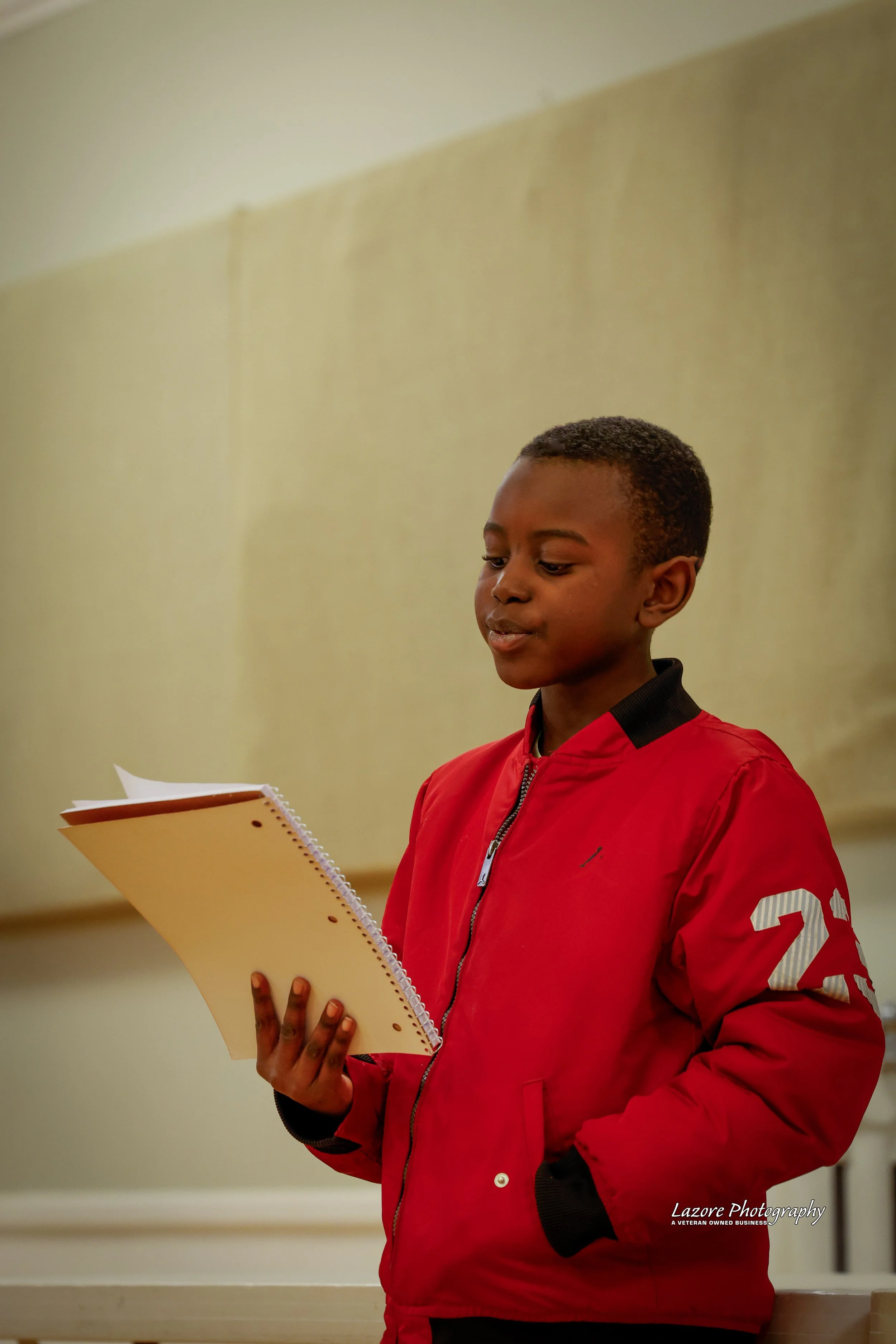 A young boy in a red jacket reading from a notepad.