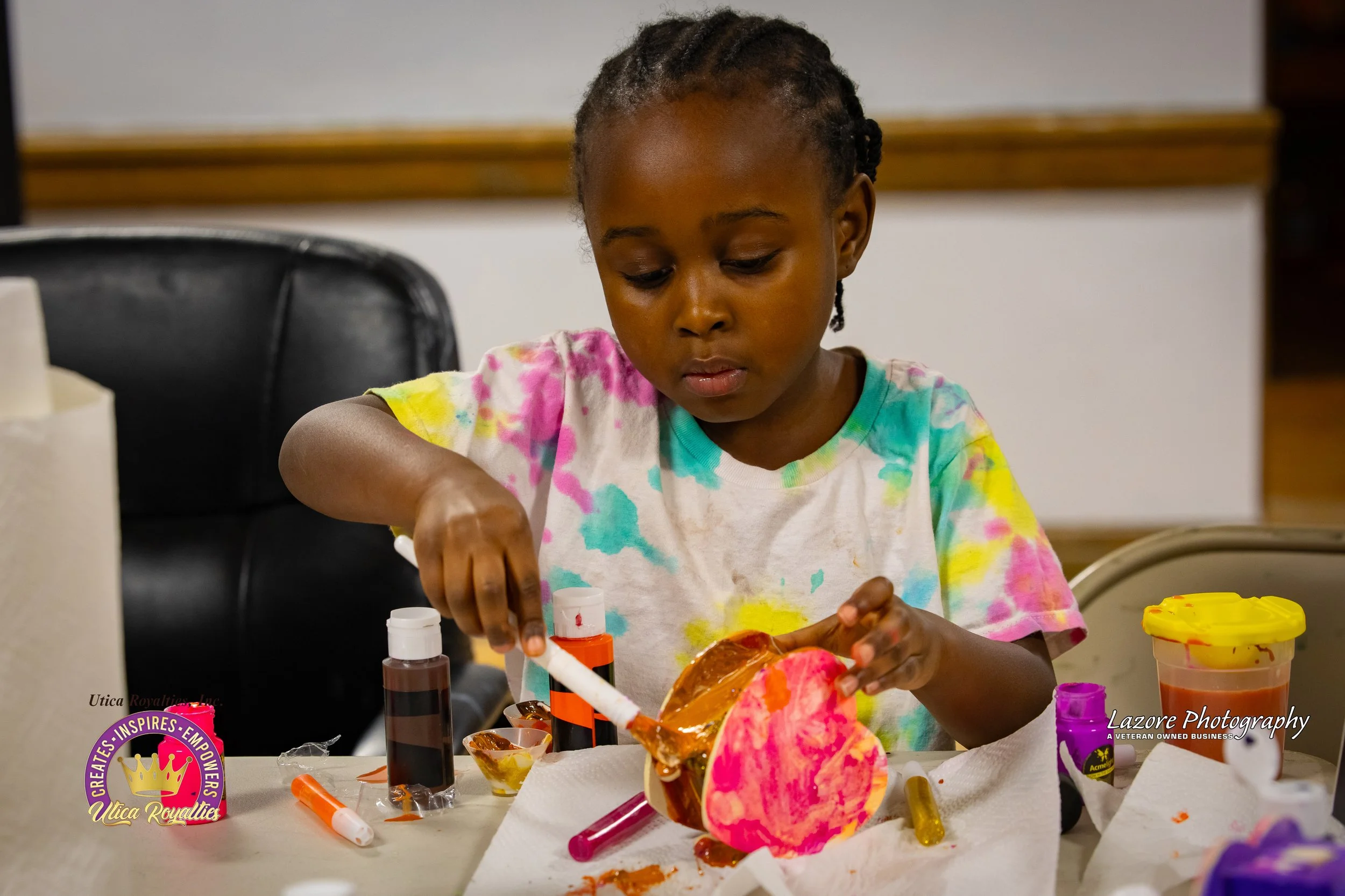 A young girl with braided hair and tie-dye shirt decorating a pumpkin with paint and glue at a table.