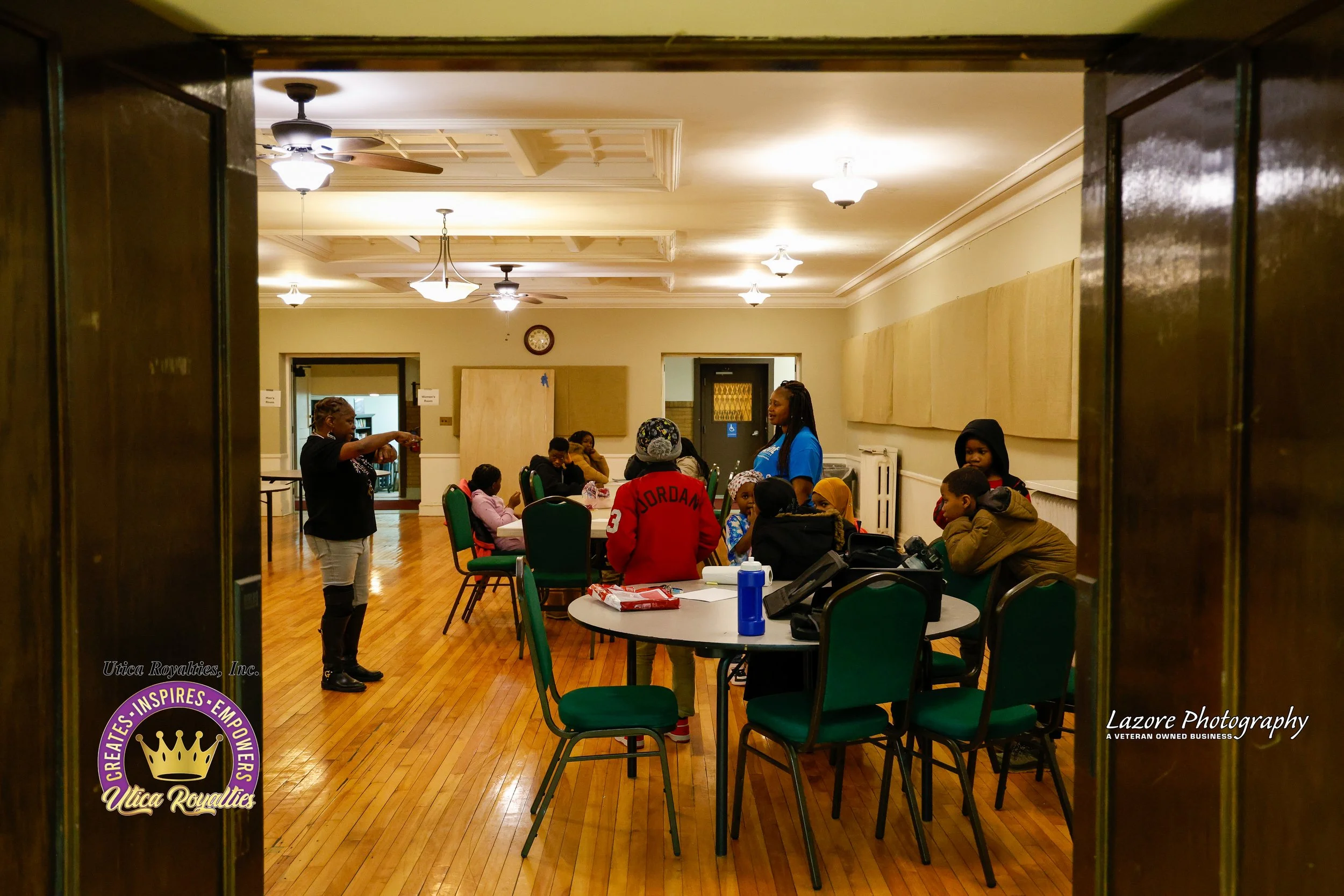 Group of children and adults in a community room engaged in an activity, with a woman standing and instructing, seen through a doorway with wooden framing.