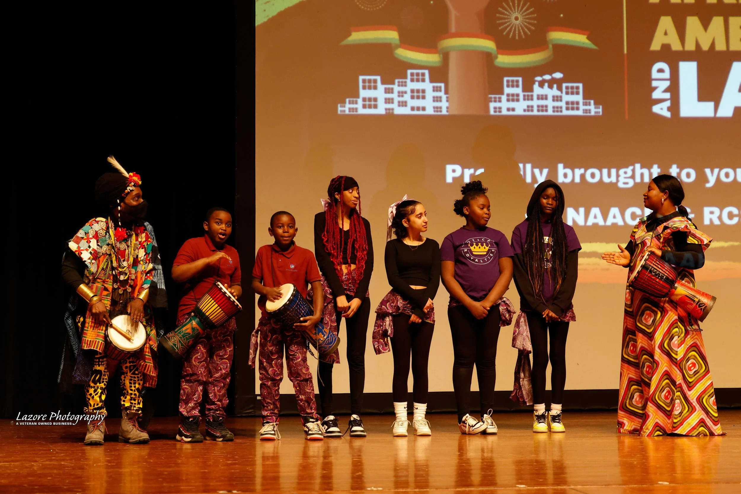Group of children and two adults on stage during a cultural presentation. The adults are wearing traditional African attire, and some children are holding drums. The background features a presentation slide with text and artwork related to African cu