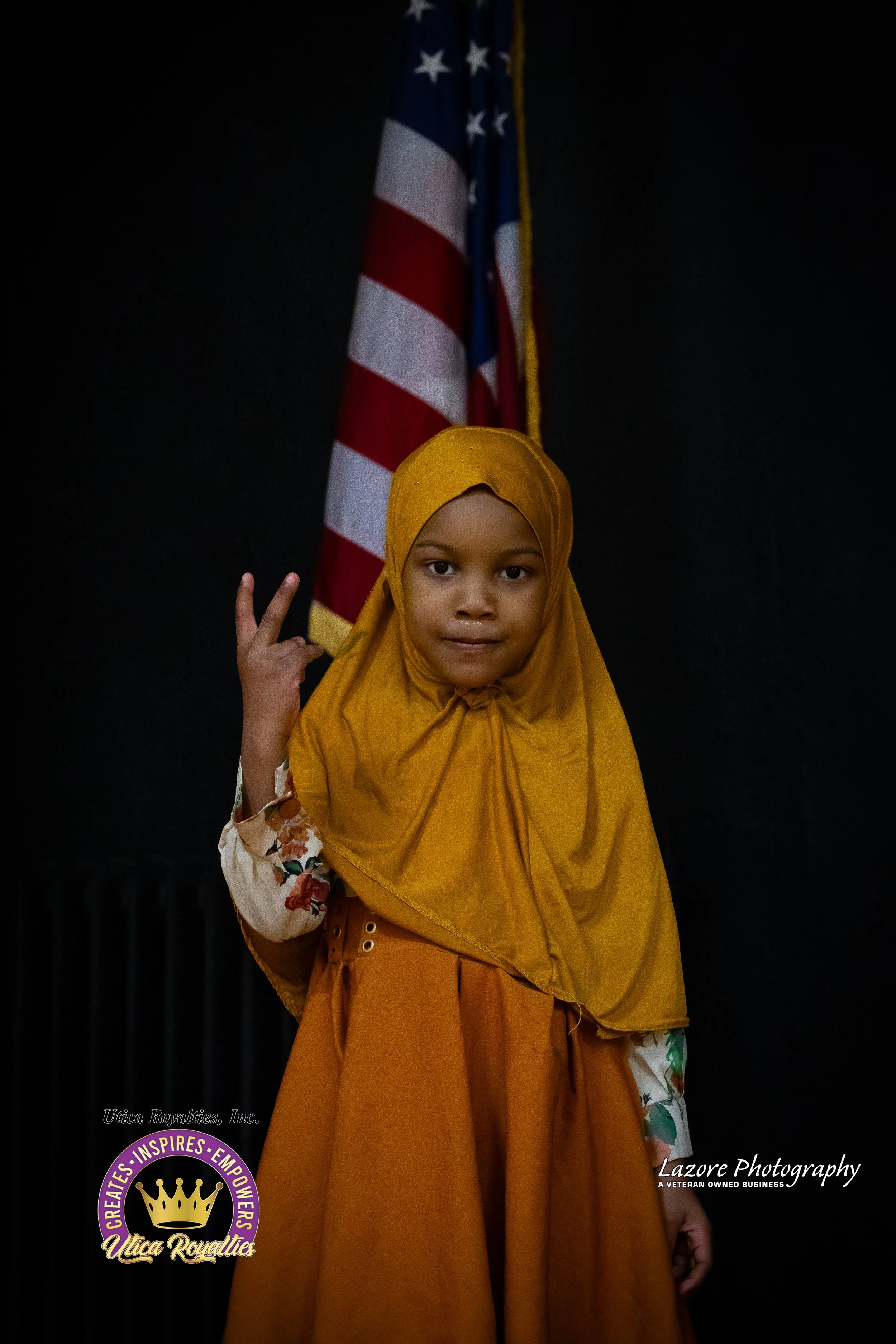 A young girl wearing an orange dress and a matching headscarf, making a peace sign with her right hand, standing in front of an American flag.