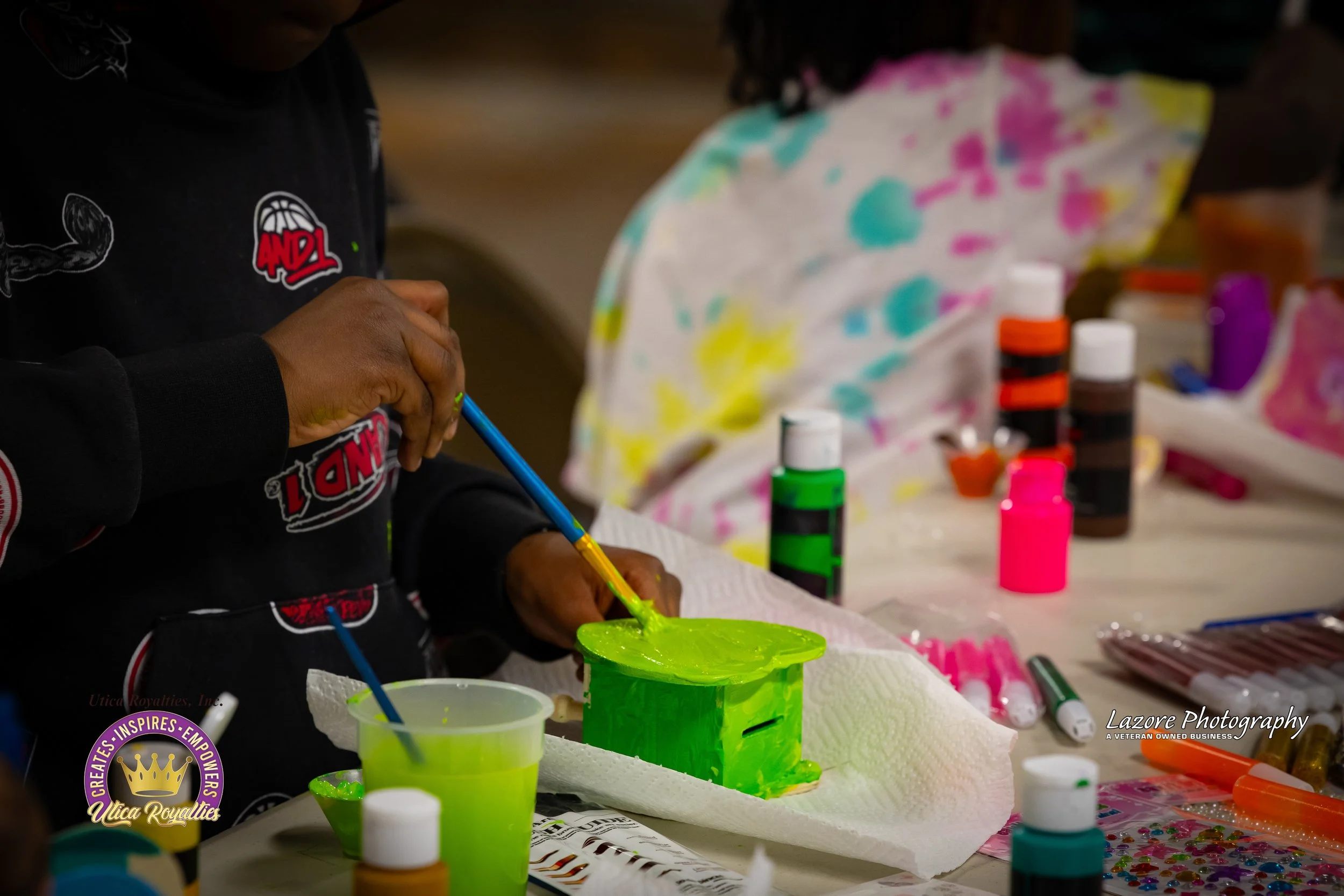 Person painting a small green wooden box with a paintbrush, surrounded by various colorful bottles of paint and craft supplies on a table.