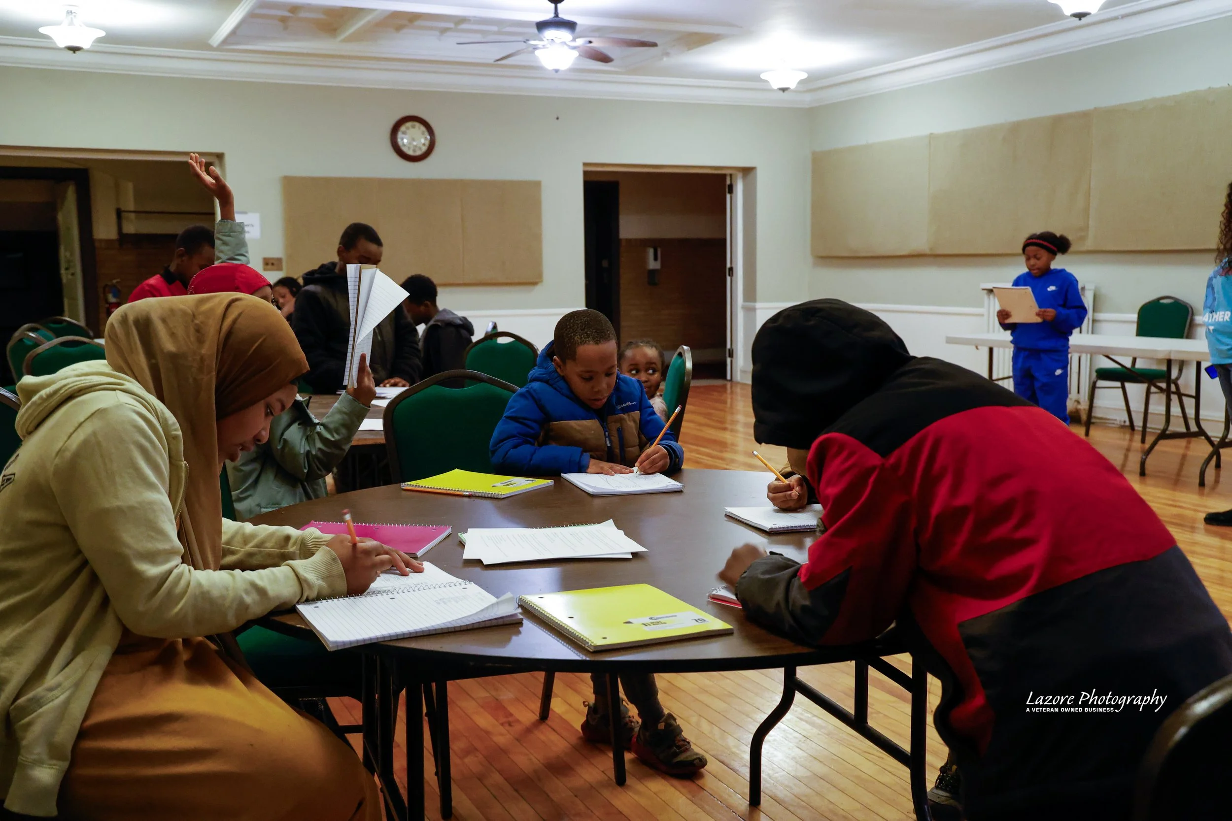 Children and adults sitting at a table in a room, engaging in reading or writing activities, with some standing in the background.