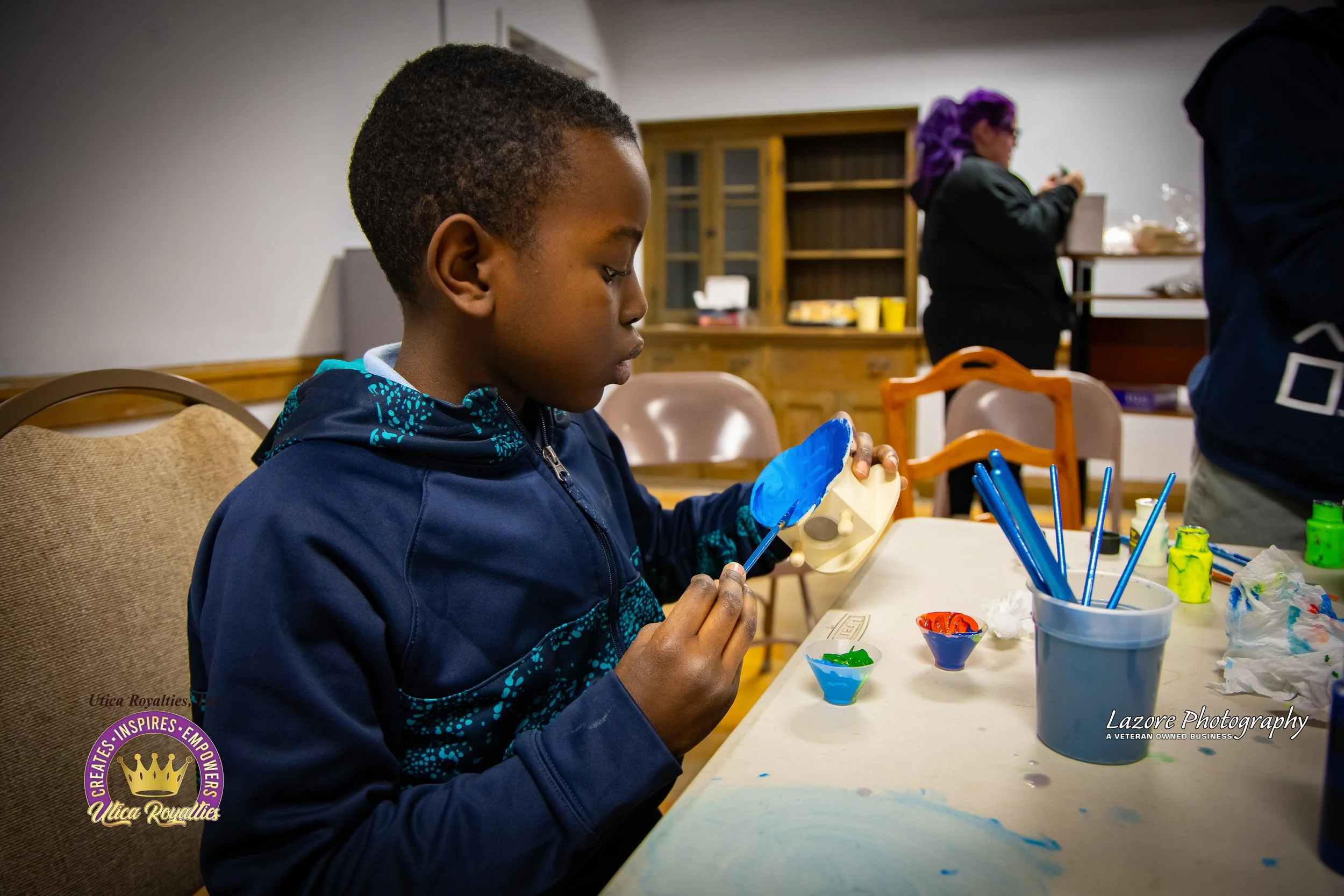 A young boy painting a ceramic mask with blue and yellow paint at a table, with paint brushes and small bowls of paint around him in a room with wooden shelves and other people in the background.
