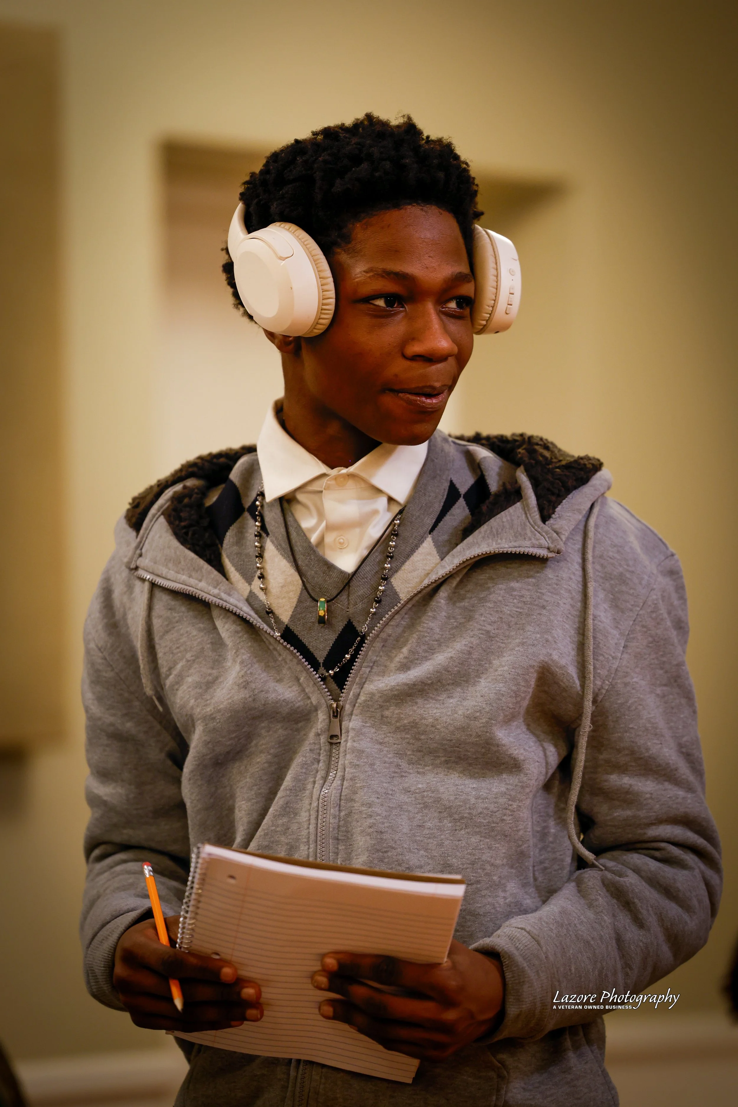 A young person with short curly hair wearing headphones, a grey hoodie over a argyle sweater, a collared shirt, and holding a notepad with a pencil.
