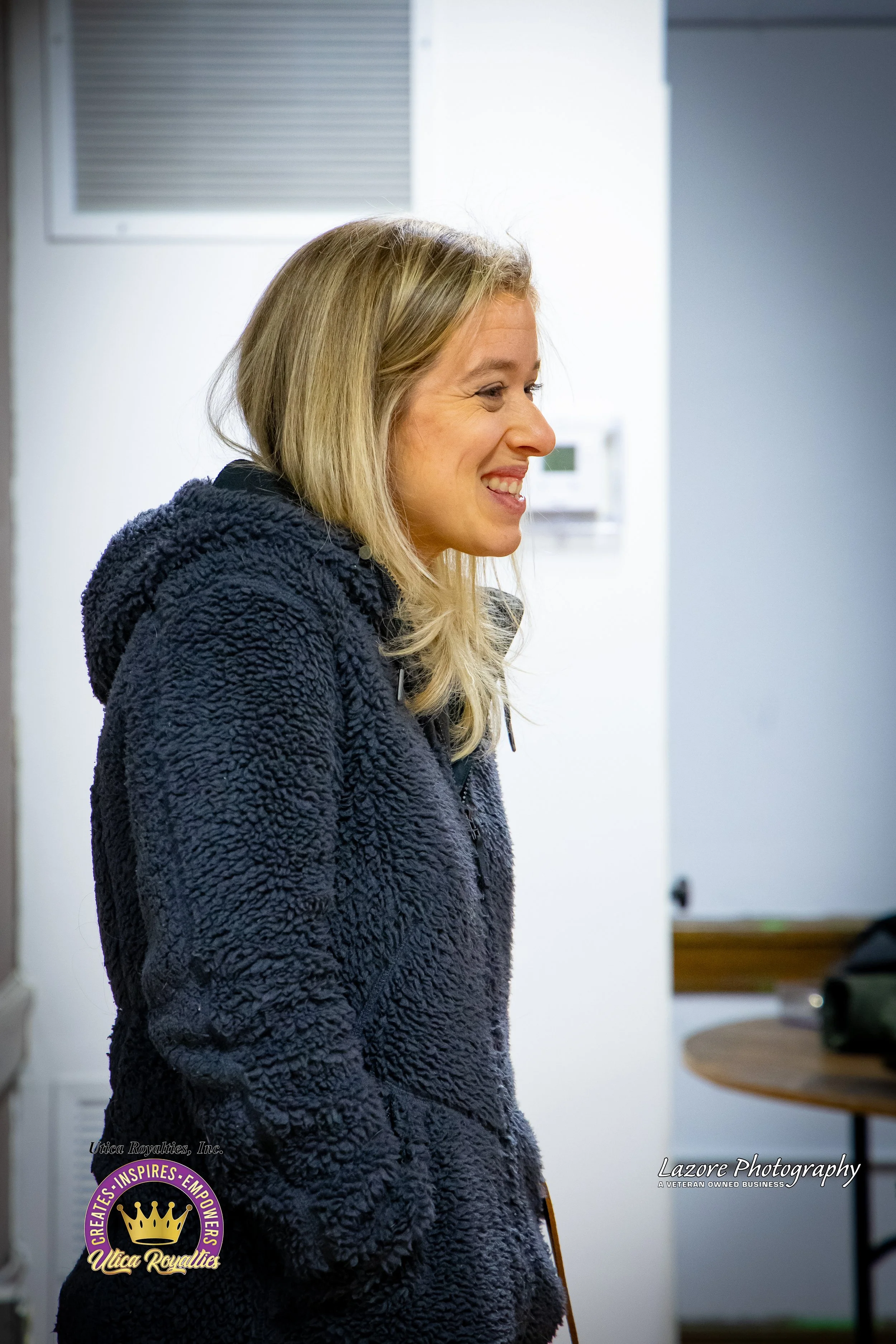A woman with blonde hair smiling, wearing a dark textured fleece jacket, standing indoors against a white wall.