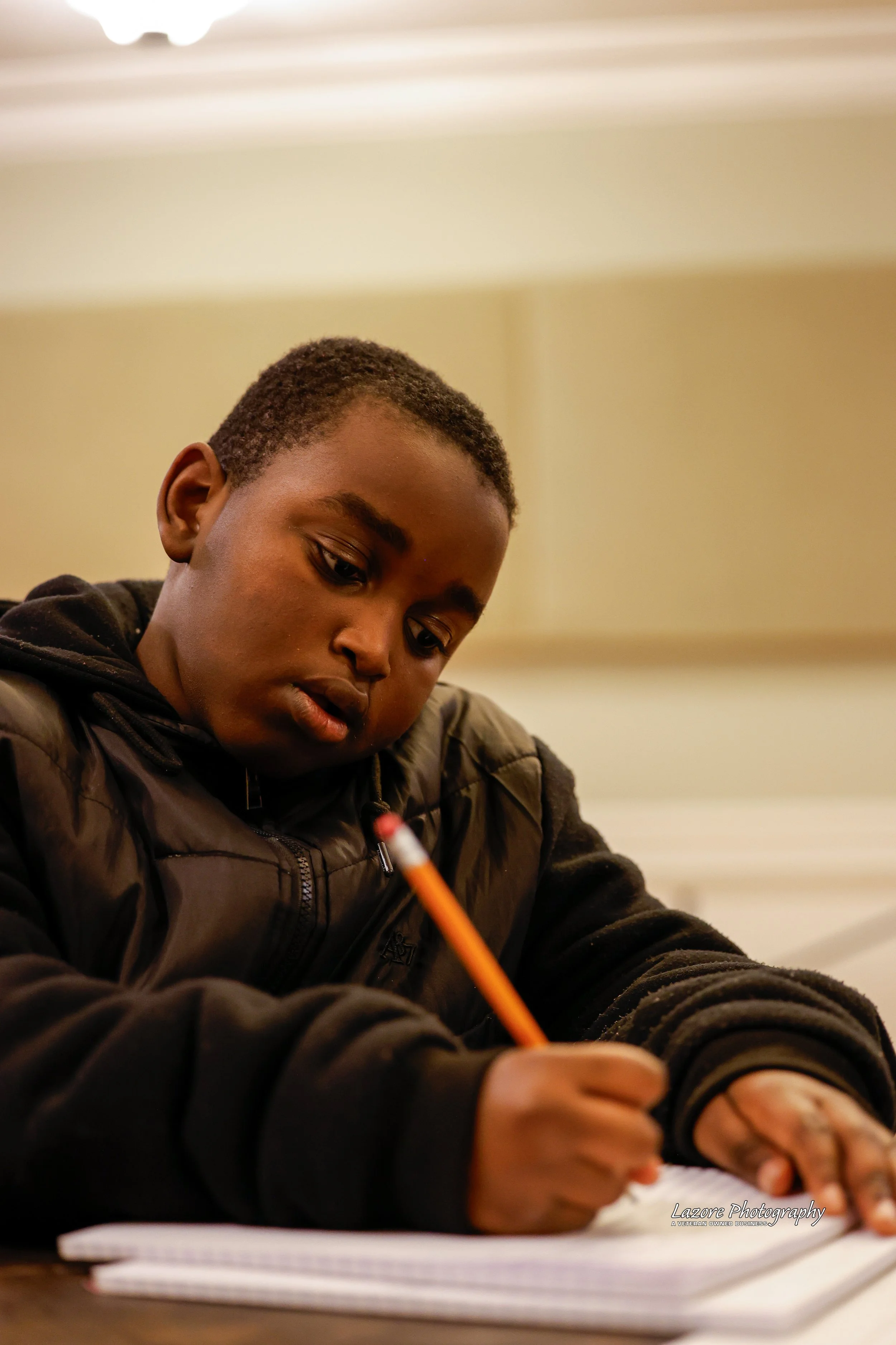 A young boy with short curly hair wearing a black jacket, sitting at a desk, writing with a pencil on paper, in a well-lit room.