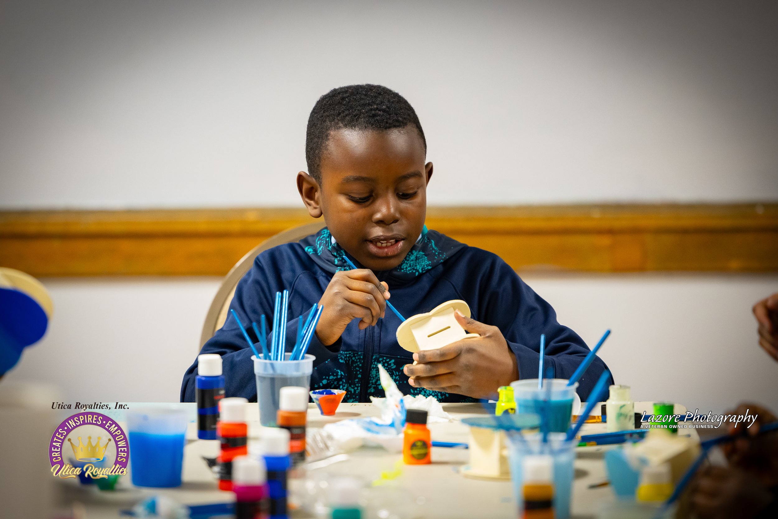 Young boy with short black hair, wearing a blue jacket, painting or decorating a small wooden box at a table with various craft supplies around him.