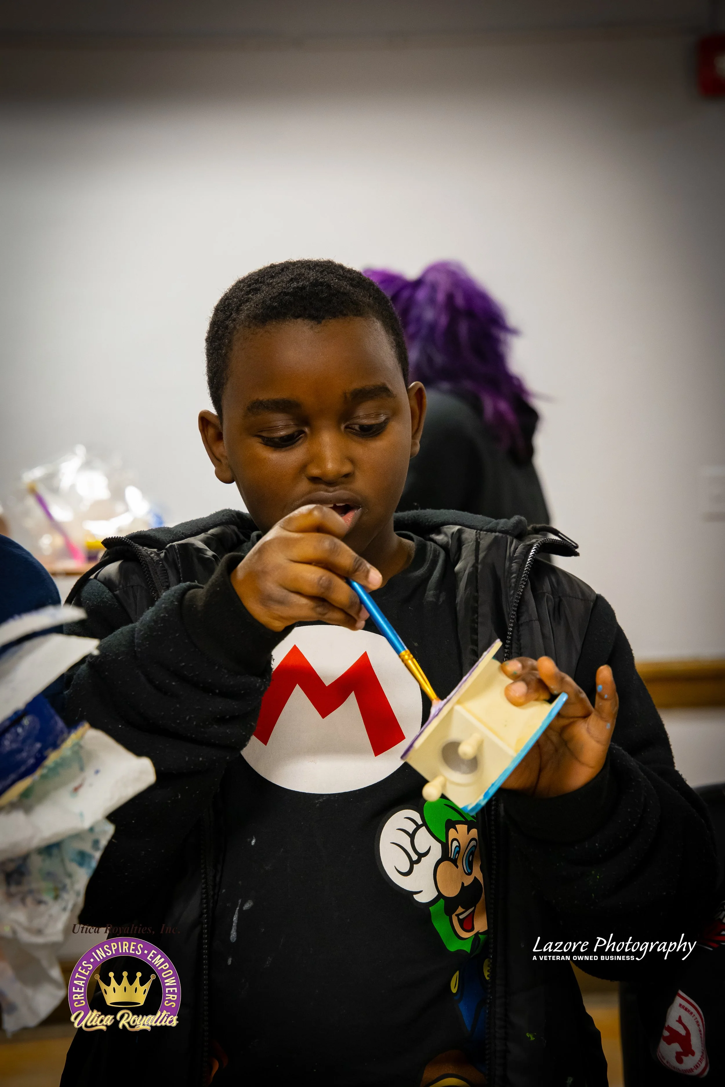 A boy with short hair painting a small wooden object with a blue and yellow brush, wearing a black hoodie over a Mario t-shirt, in an indoor setting.