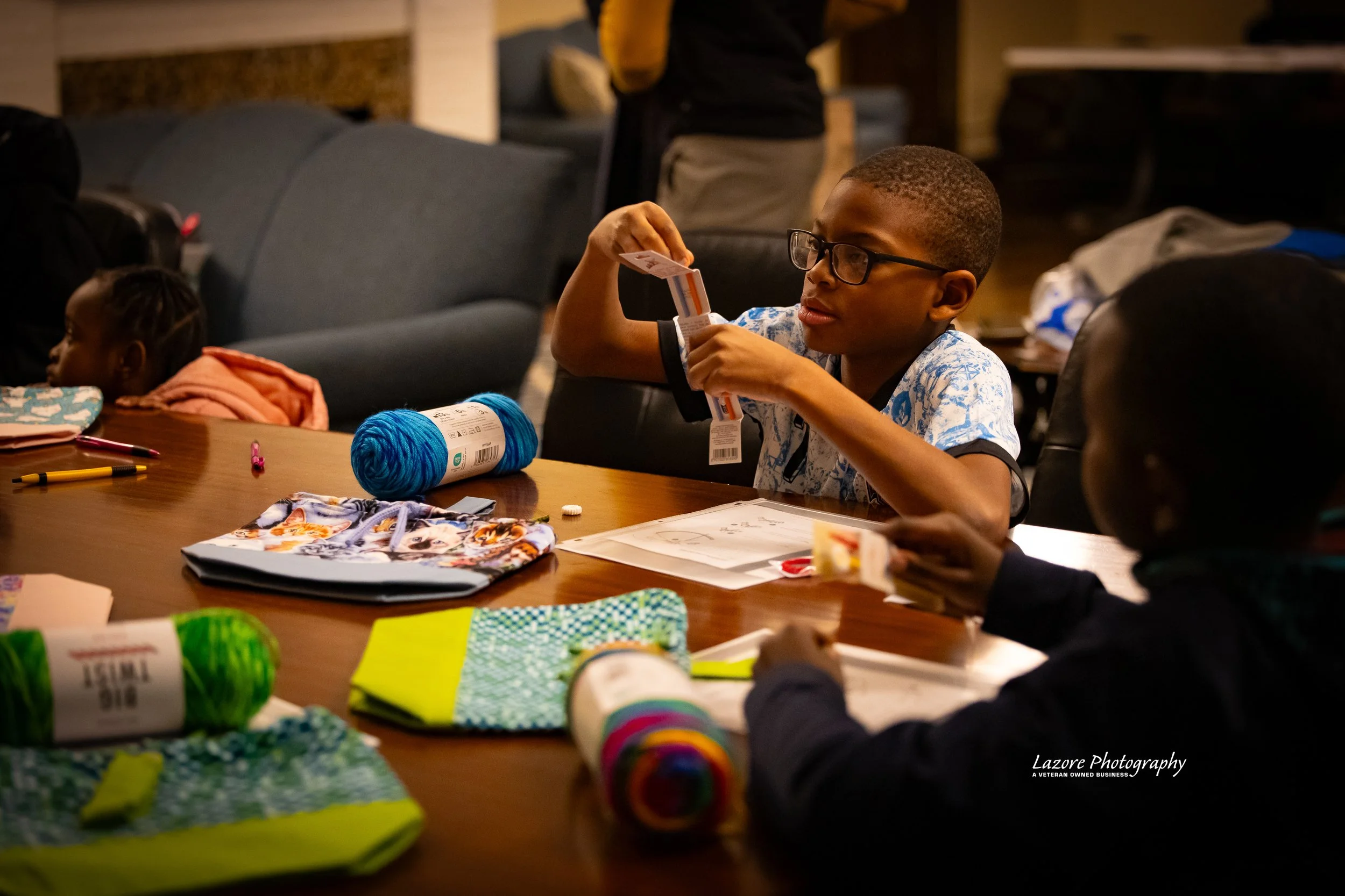 Young boy sitting at a table, wrapping a gift with wrapping paper, surrounded by yarn, markers, and fabric in a cozy indoor setting.