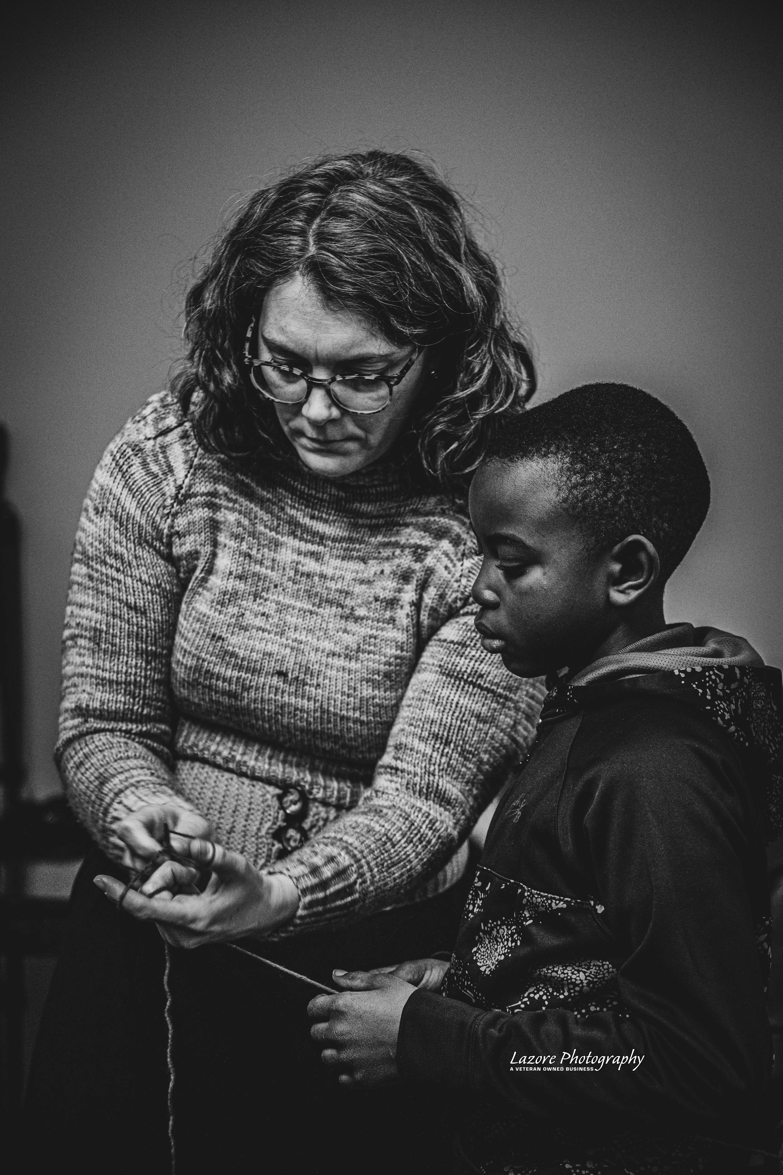 A woman with glasses and curly hair teaching knitting to a young boy in black and white.