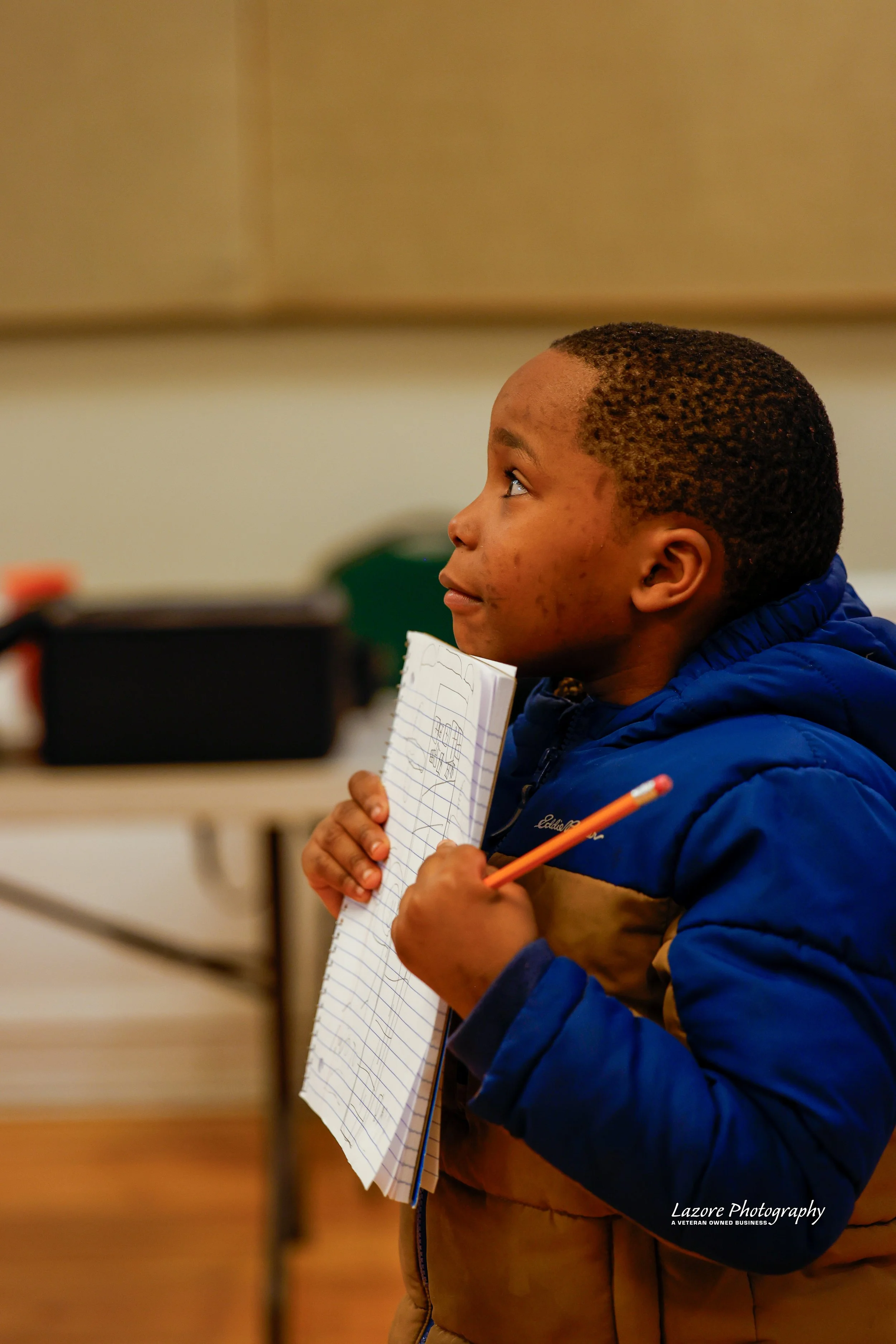 Young boy in blue and brown jacket holding a lined notebook and pencil, attentively listening in a classroom setting.