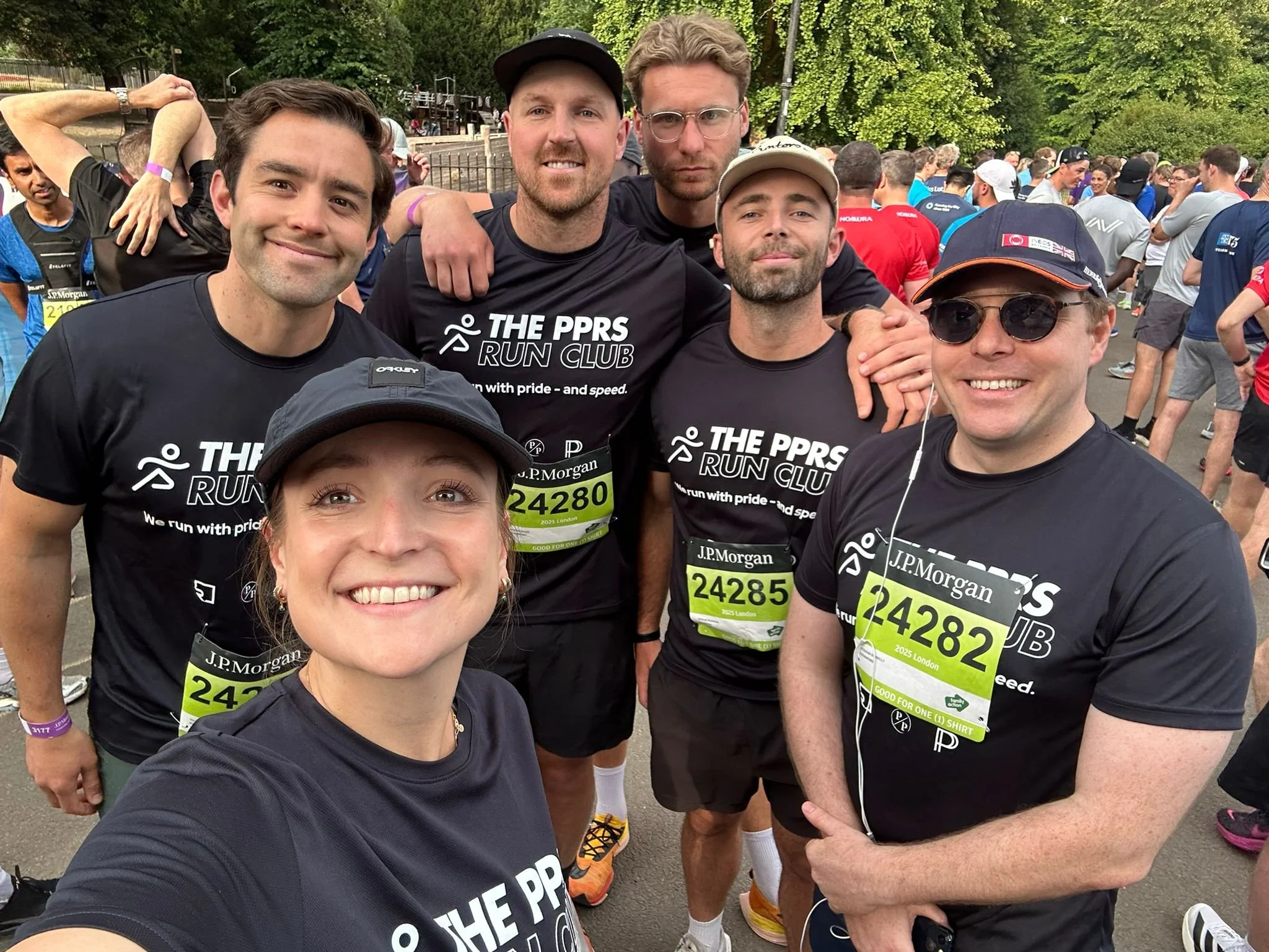 Group of smiling runners at a marathon event, wearing matching black T-shirts with the logo and text "THE PPRS RUN CLUB," with a crowd of other runners behind them at an outdoor park setting.