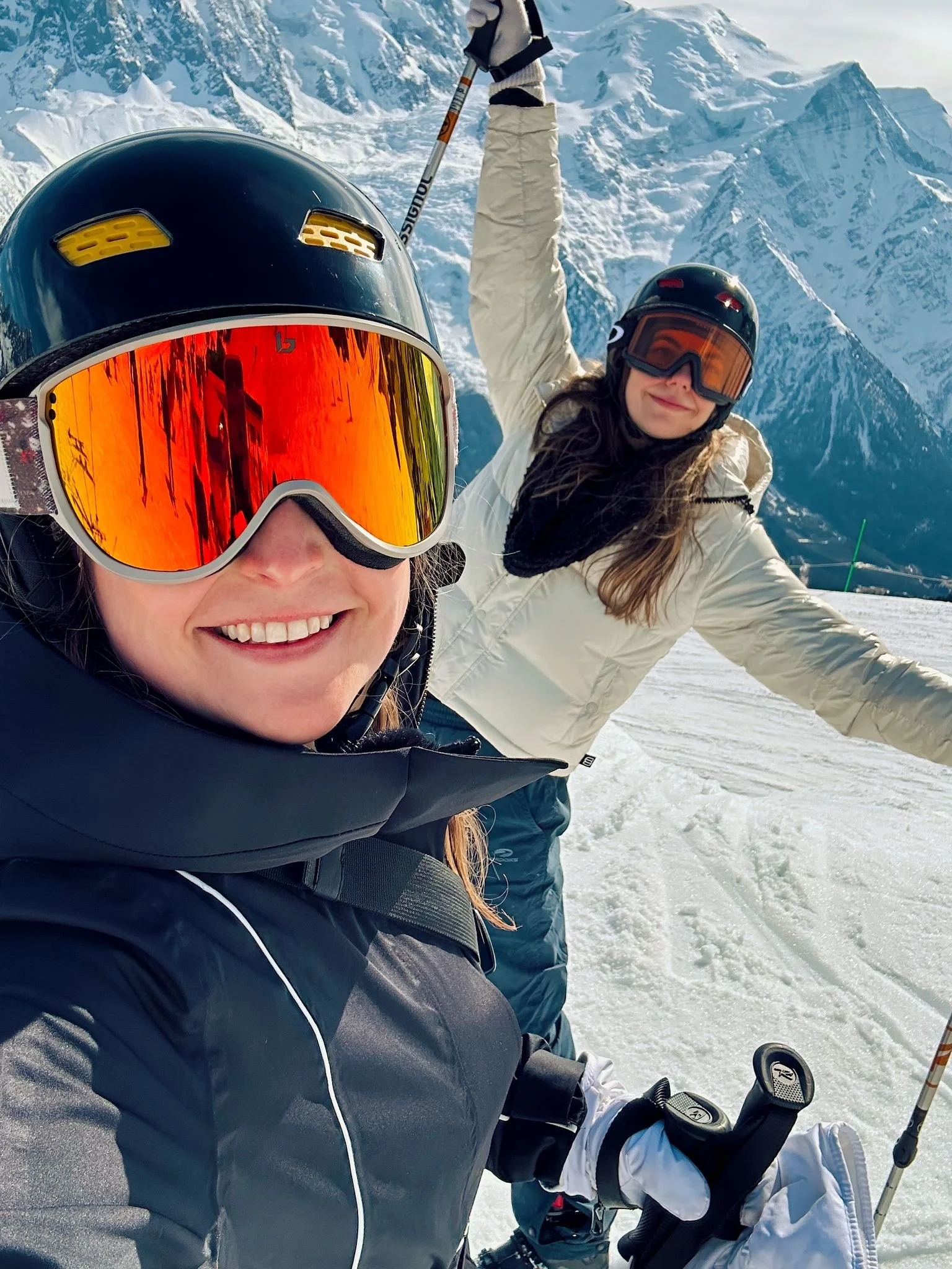 Two women in ski gear taking a selfie on a snowy mountain with snow-covered peaks in the background.