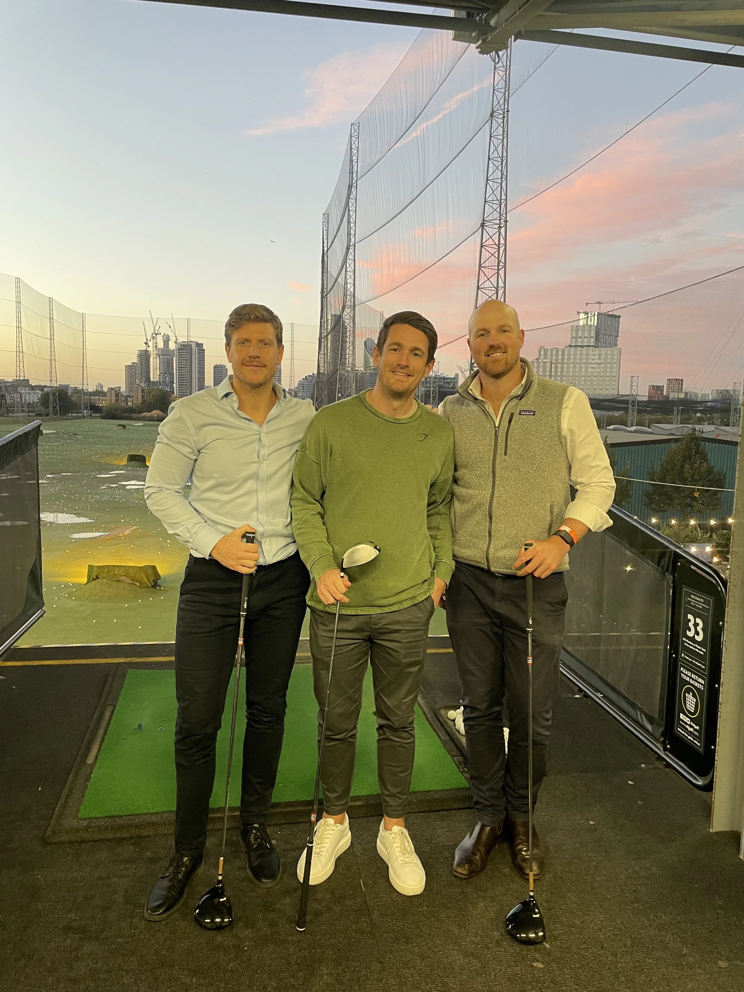 Three men standing on a golf driving range at sunset, holding golf clubs, with city skyline and tall buildings in the background.