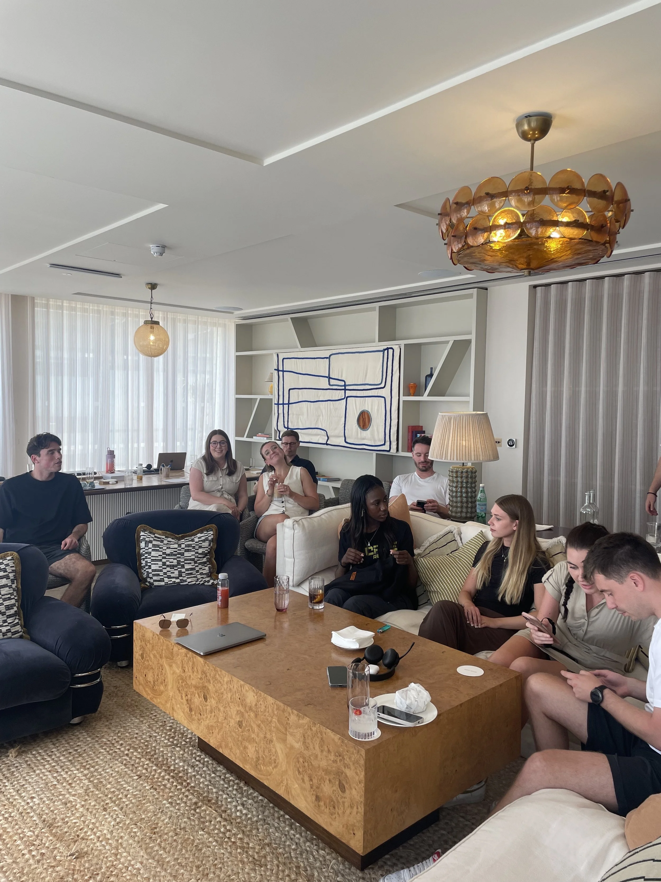 Group of people sitting and standing in a modern living room with a large wooden coffee table, artwork on the wall, and various items like drinks and electronics on the table.