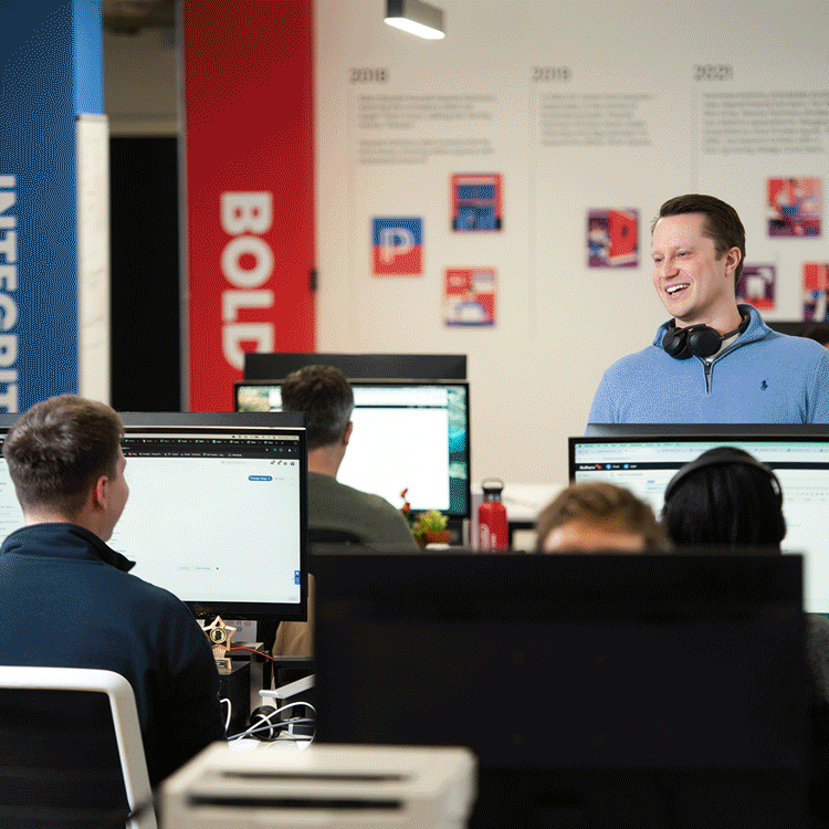 A young man with headphones around his neck is smiling and speaking to a group of people in a computer classroom or office setting.